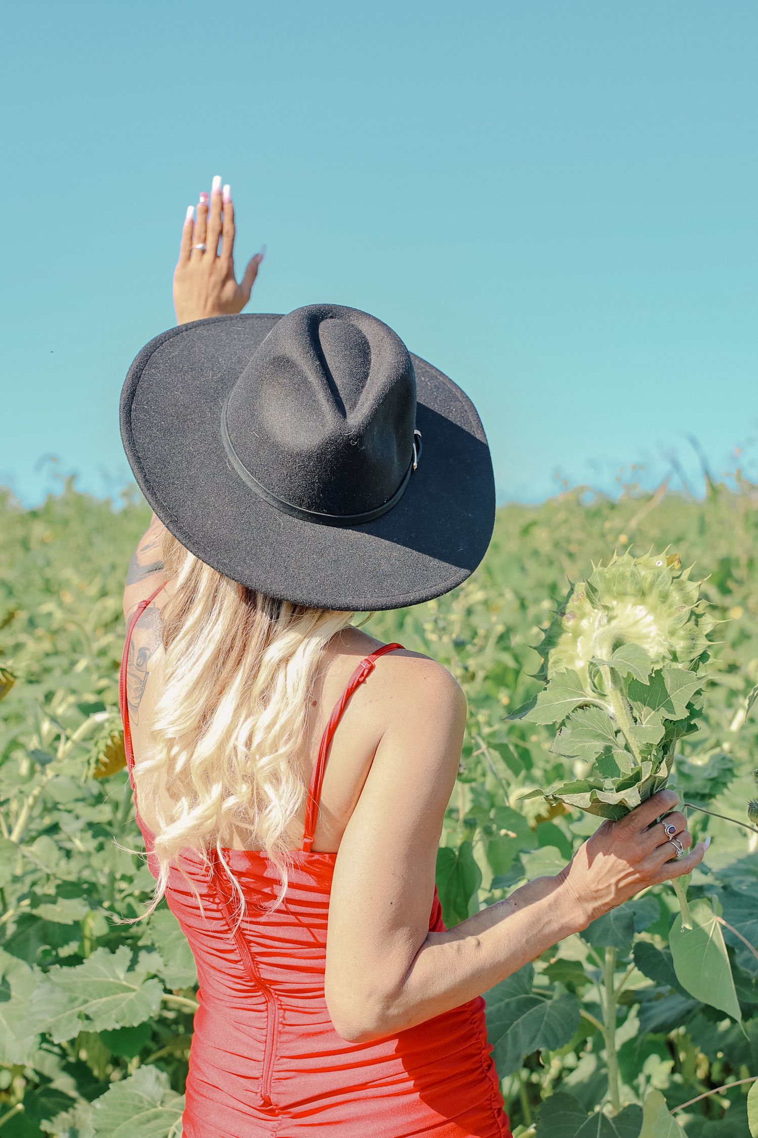 Woman in red dress and gray hat holds flowers while standing in a sunny field on a bright summer day.
