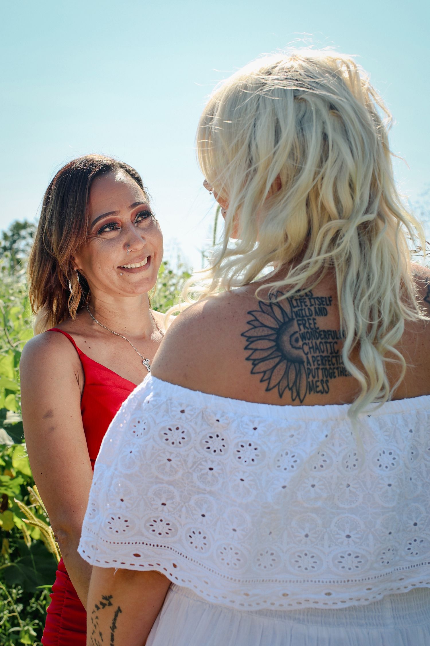 Two friends share a moment in sunny vineyard, one wearing white eyelet dress with visible sunflower tattoo.