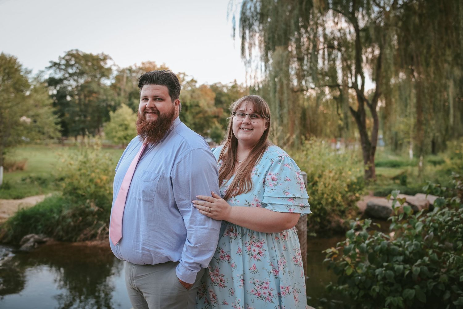 A couple stands together by a peaceful pond with willow trees and lush greenery in the background.