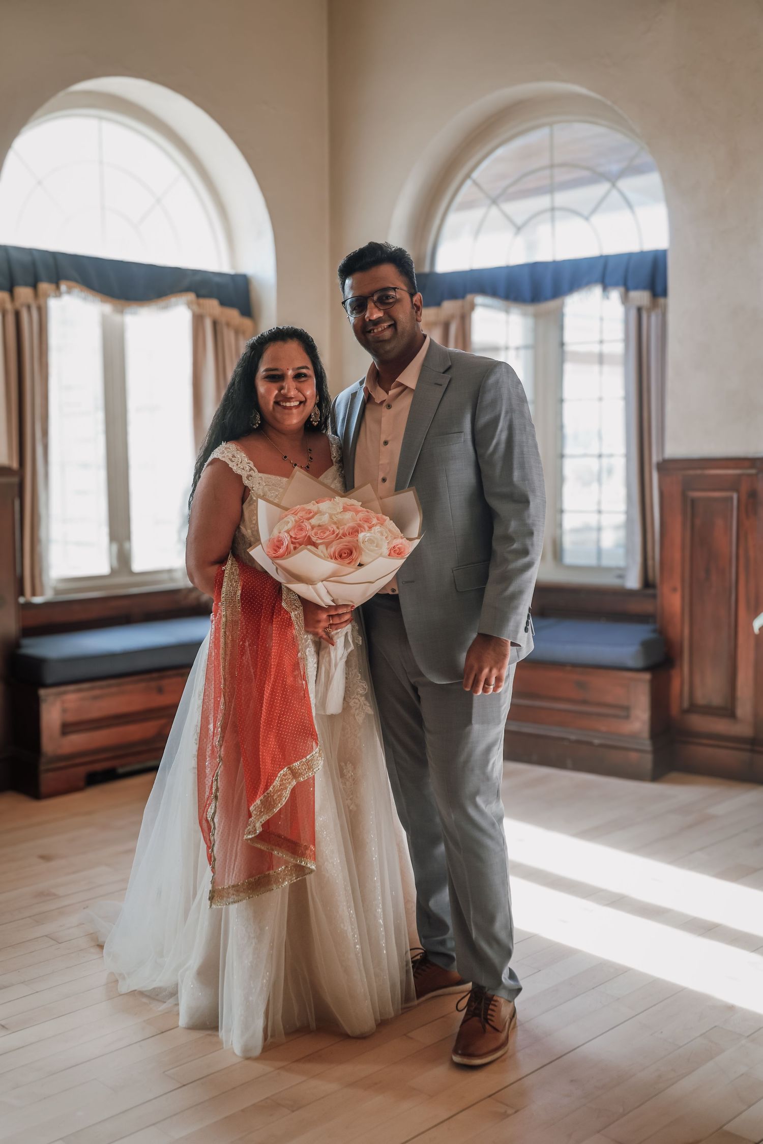 A couple in wedding attire shares a tender moment in a bright sunlit room with arched windows and wooden floors.