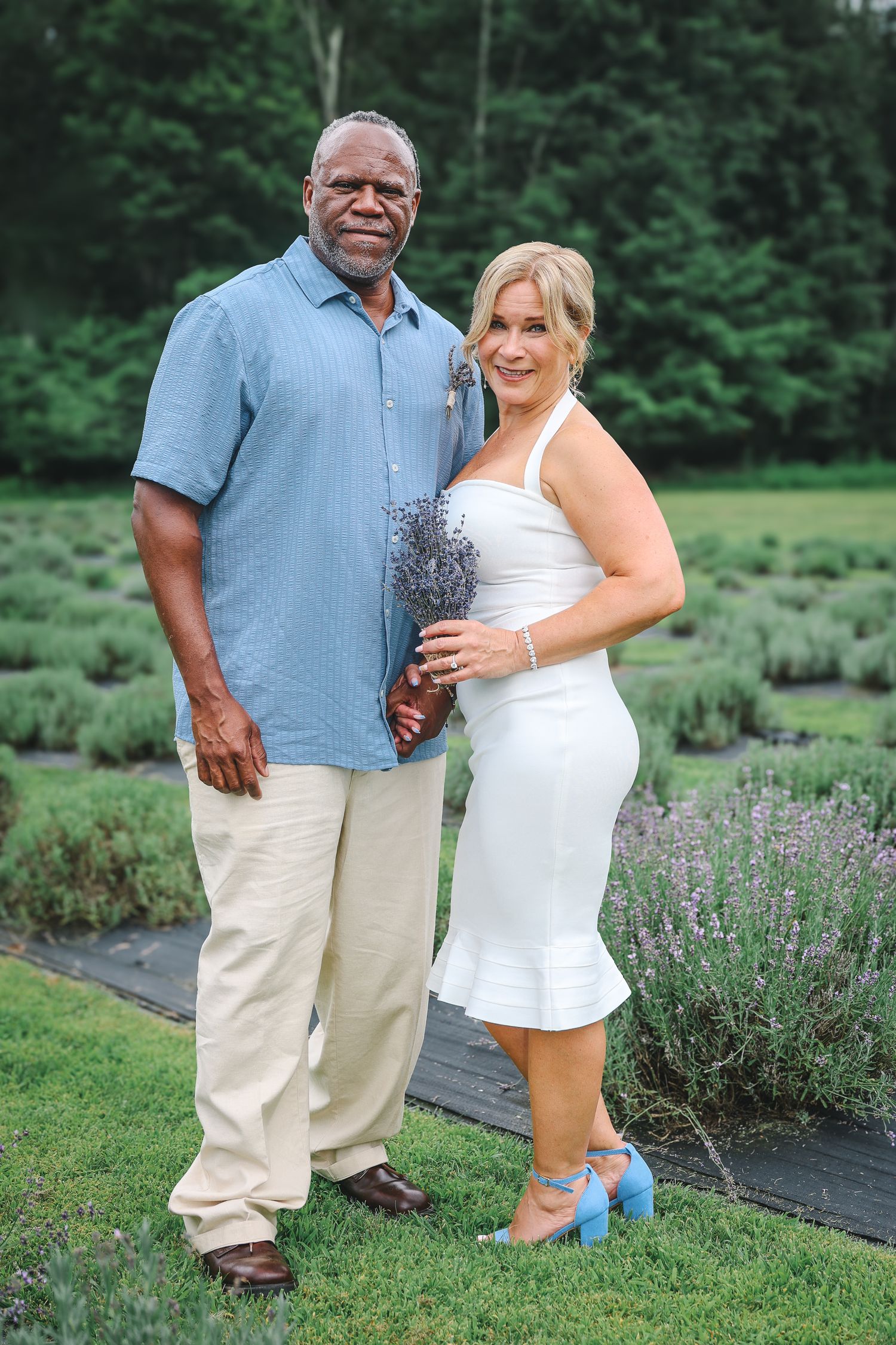 A couple poses together on green grass with lavender flowers in the background during summer.