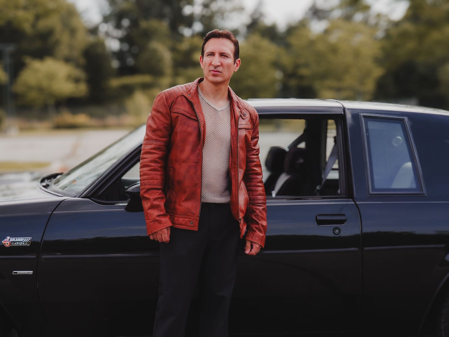 Person in red leather jacket stands beside dark colored car in outdoor setting.