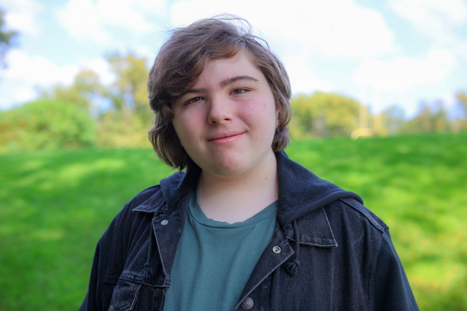 A person in a black leather jacket smiles at the camera while standing in a grassy field on a sunny day.
