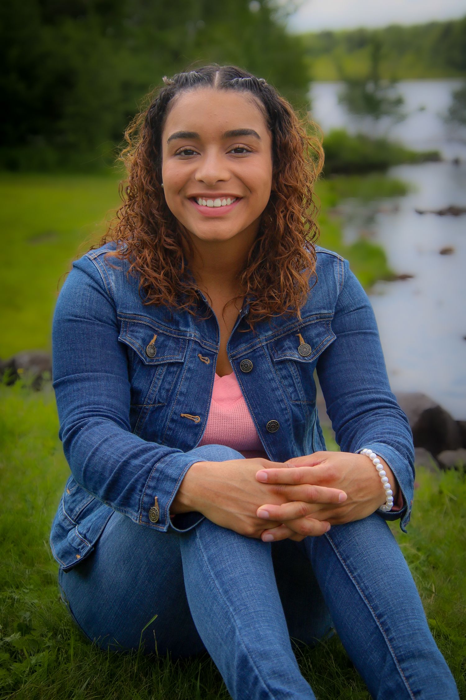 A woman in a denim jacket sits casually on grass near a lake, showcasing a warm and genuine expression.