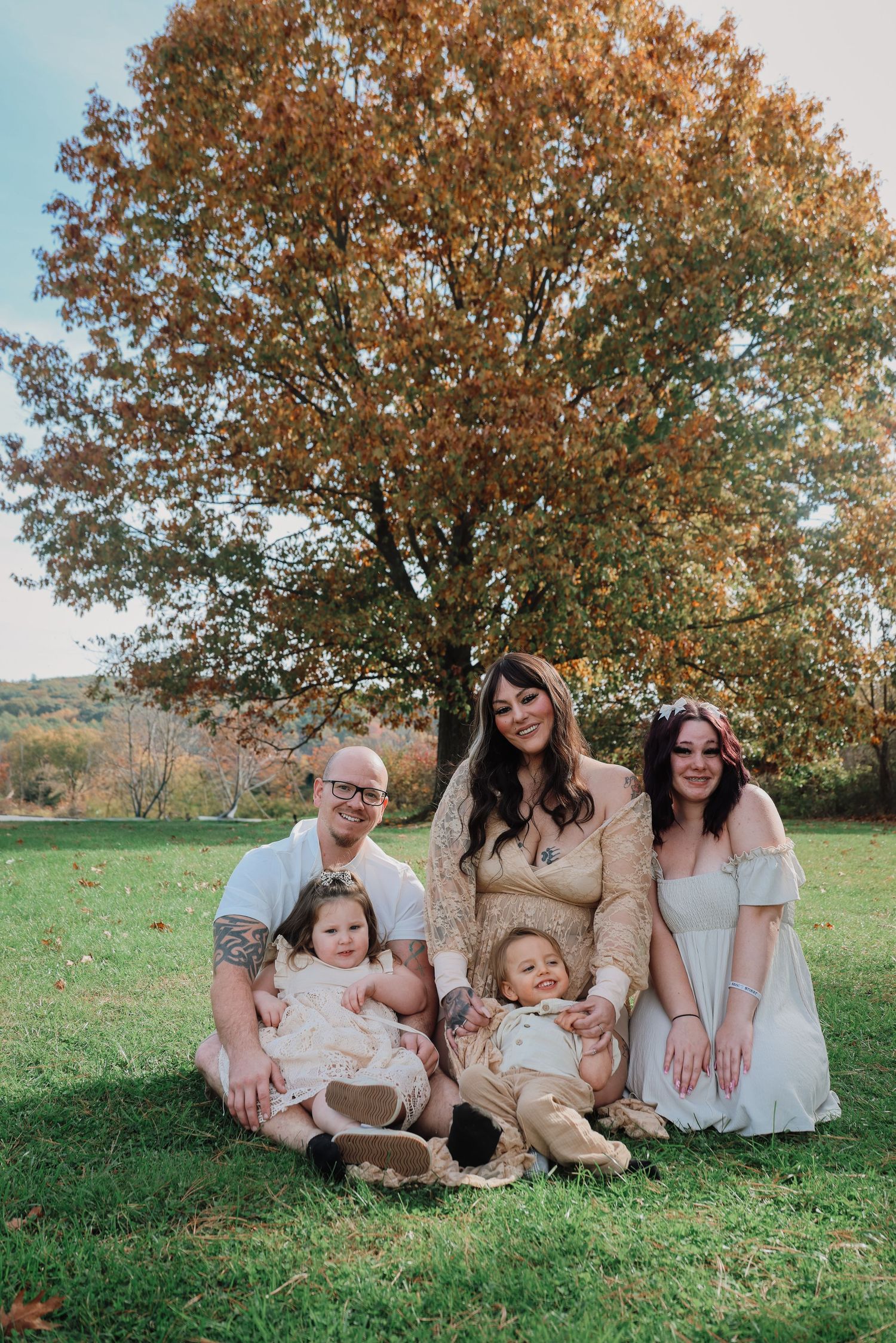 A family sits together on grassy hill during autumn with fall foliage and trees in the background.