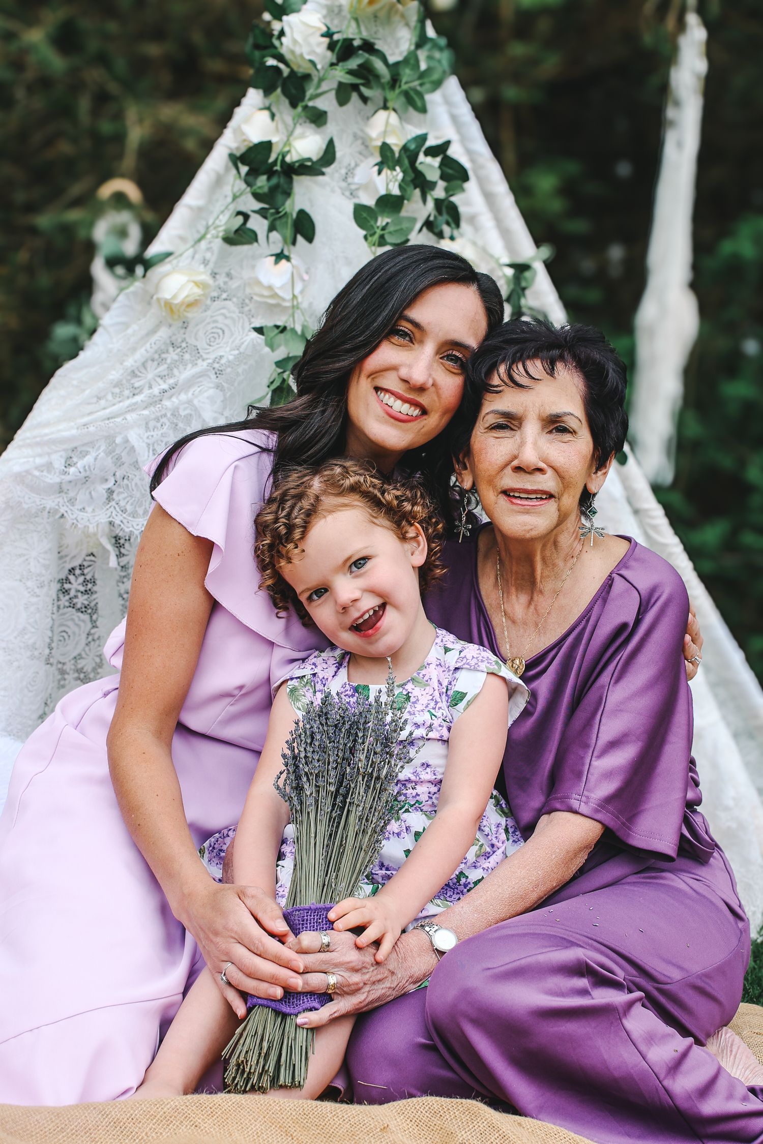 Three generations pose together in purple and pink dresses with lavender bouquet against white lace teepee backdrop.