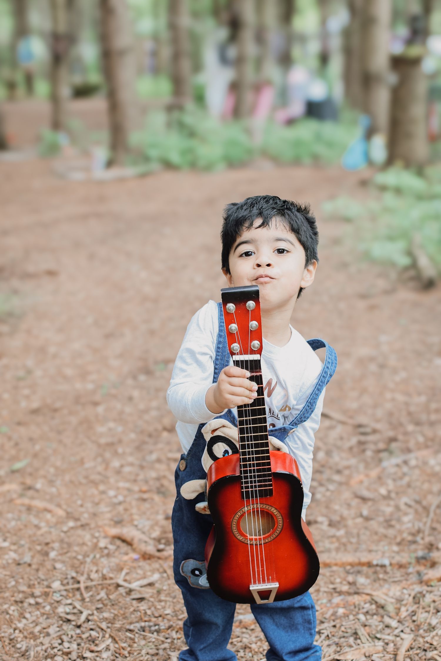 Young child in overalls holds a small acoustic guitar while standing on a dirt path in a forest setting.