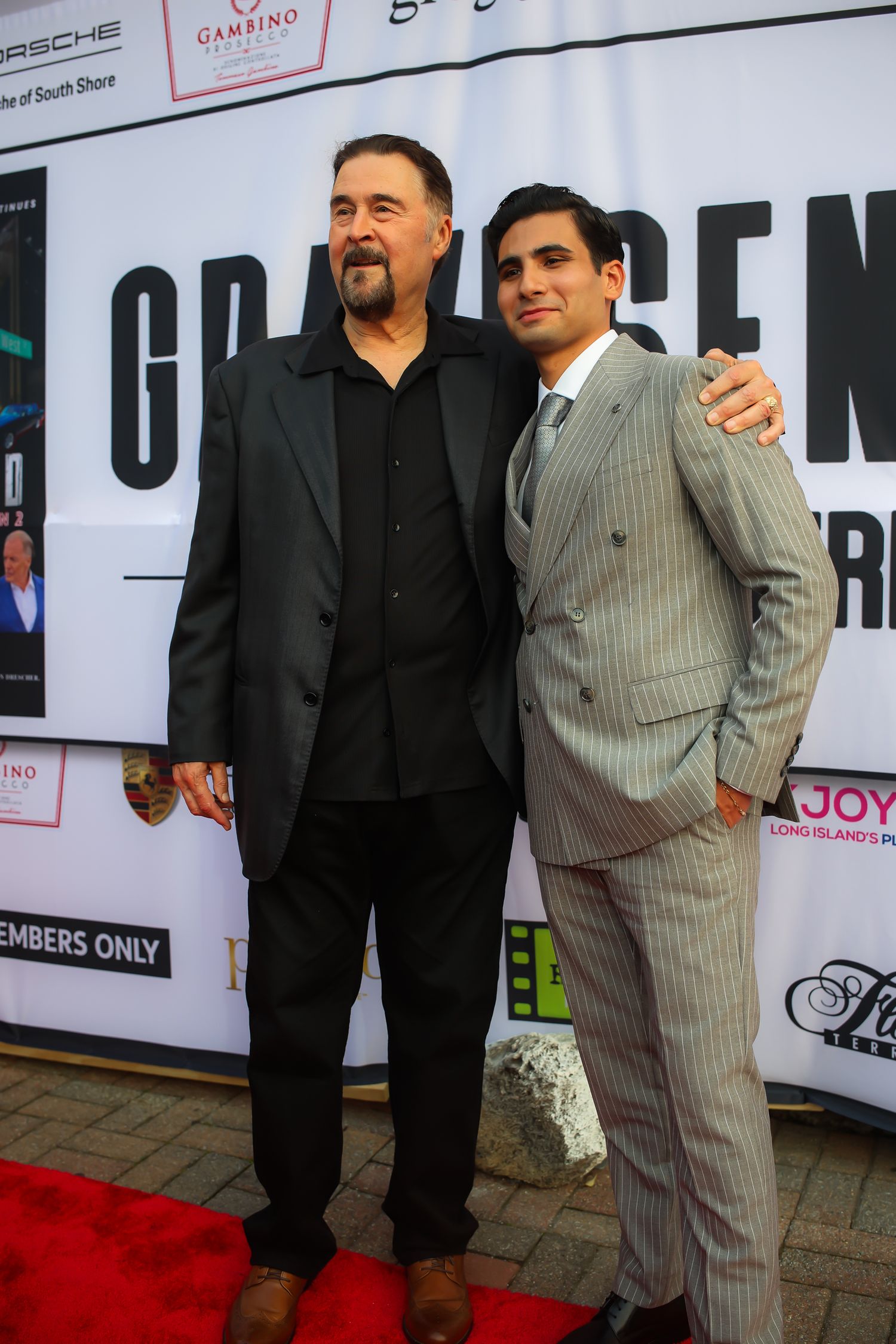 Two men pose together on a red carpet at a premiere event, one wearing all black and one in a pinstriped suit.