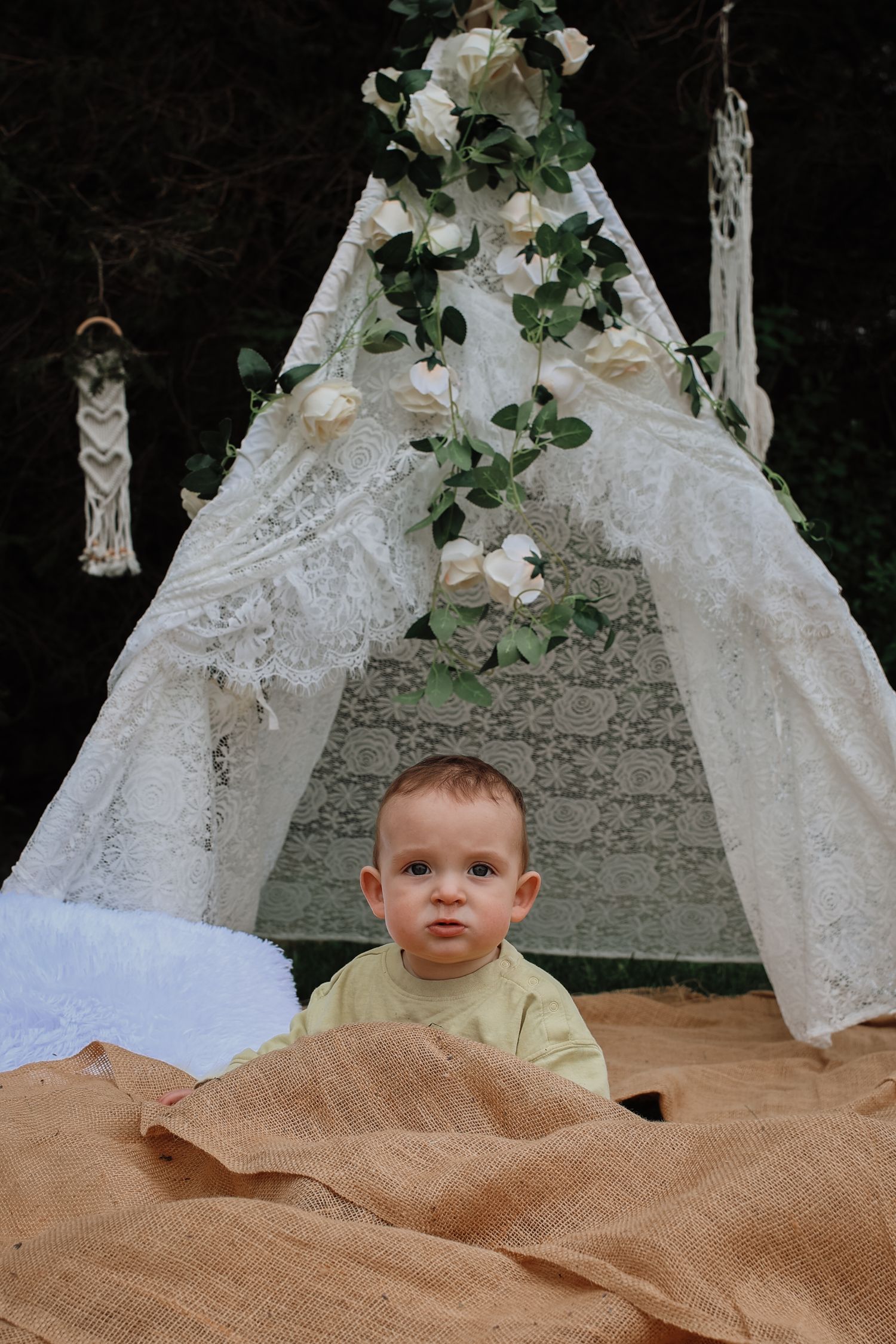 A small child sits under a white lace teepee decorated with flowers and greenery, with a burlap blanket.