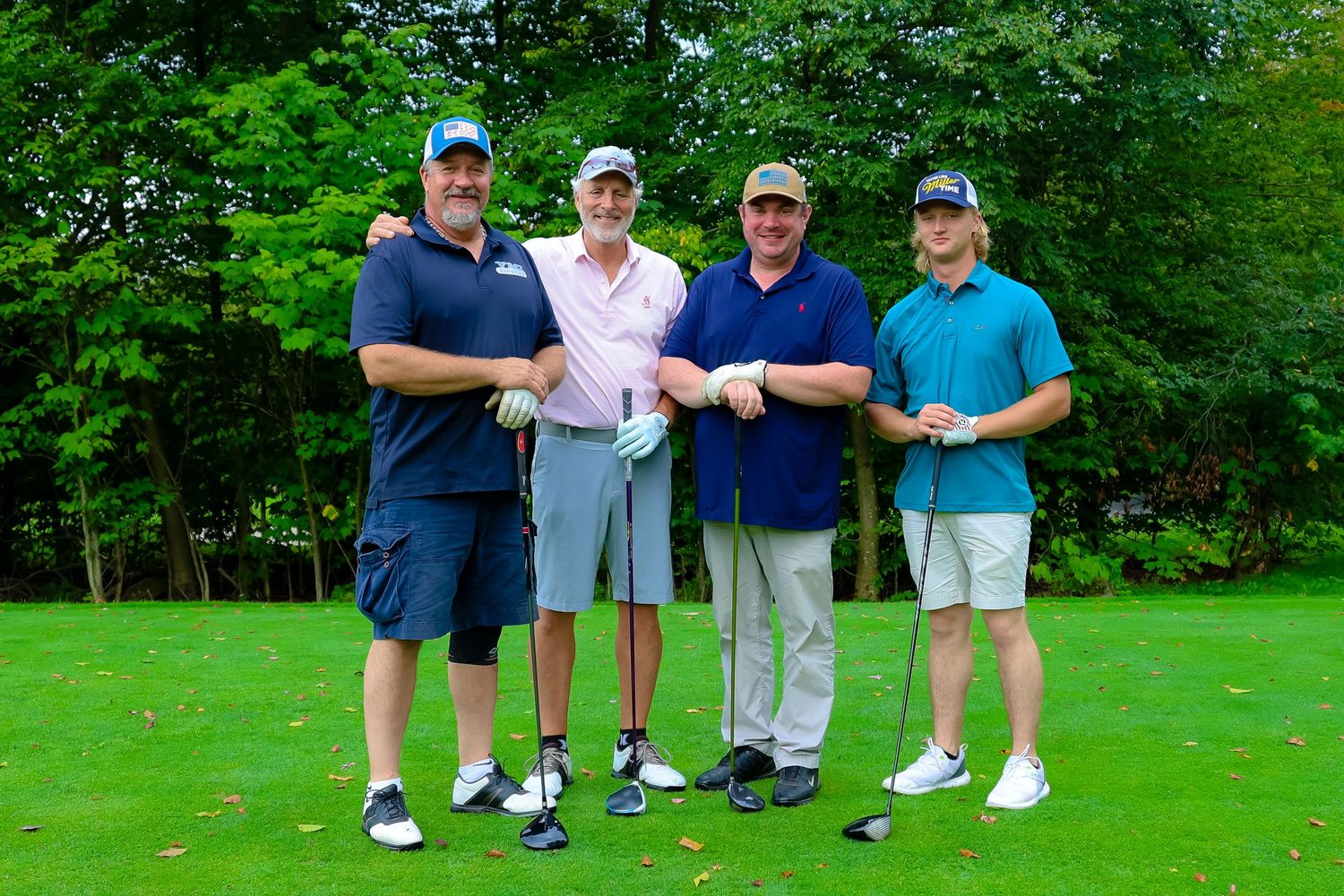 Four golfers pose together on a lush green golf course with trees in the background.