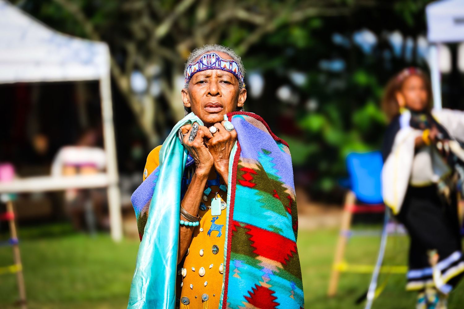 A performer in traditional Native American regalia with colorful blanket and beaded headband at an outdoor cultural event.