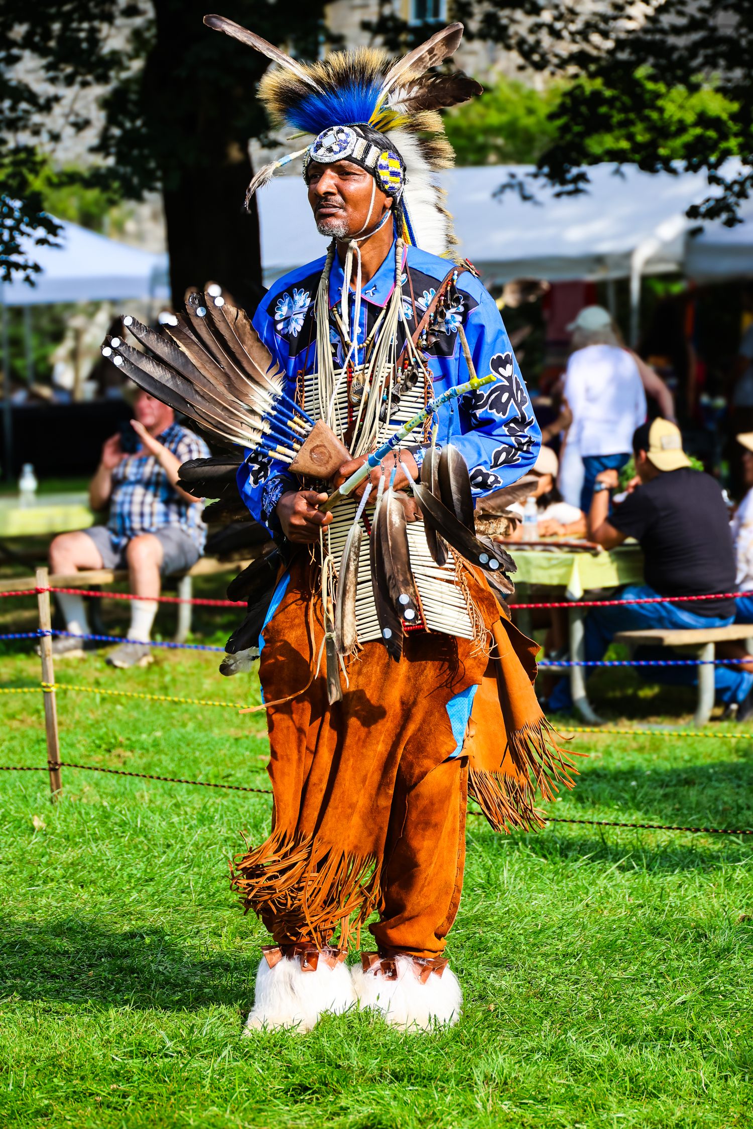 Traditional Native American dancer in vibrant blue and tan regalia performs at outdoor cultural festival.