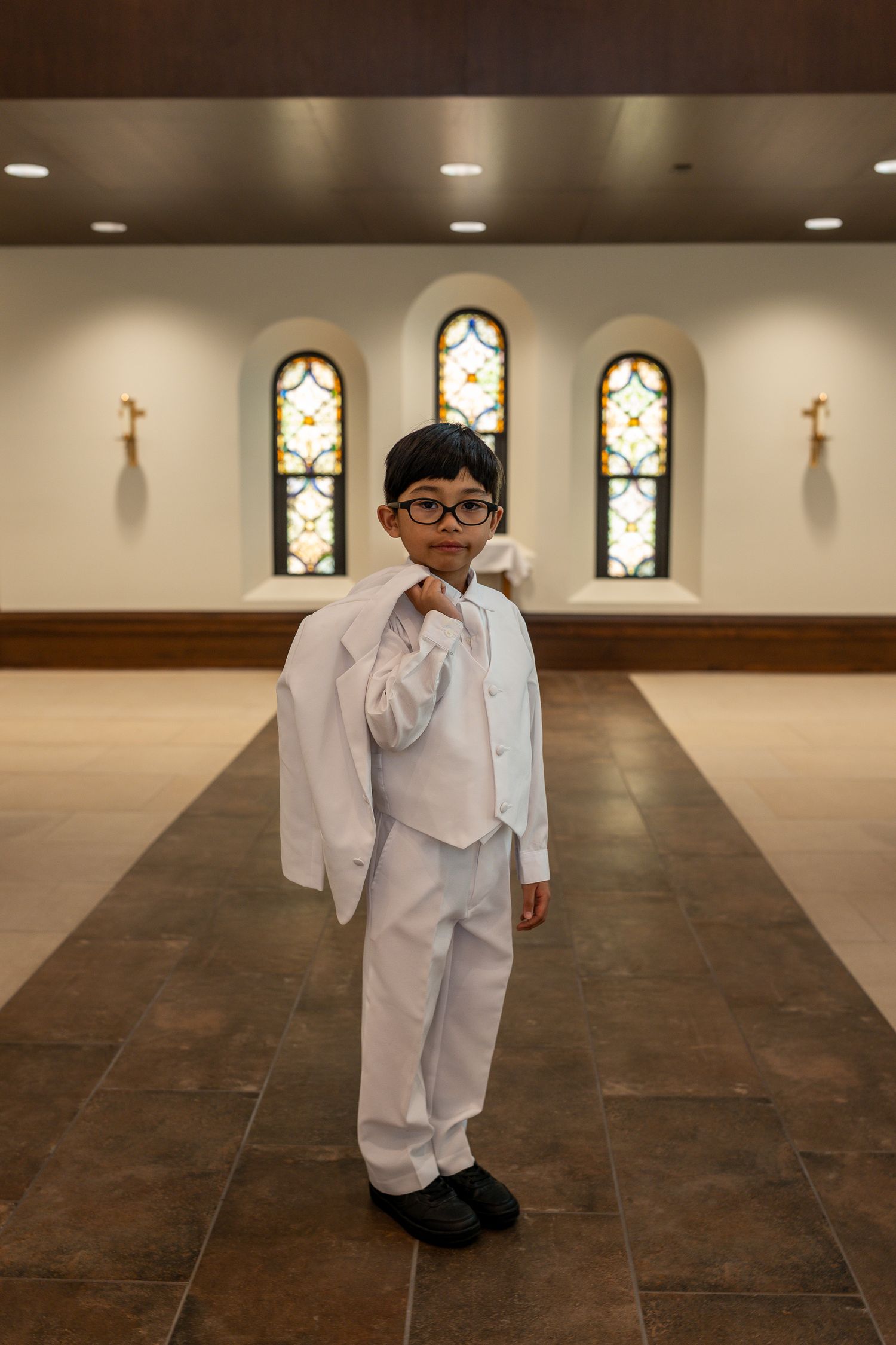 A young child in a formal white suit stands in a church with stained glass windows and crosses on the wall.