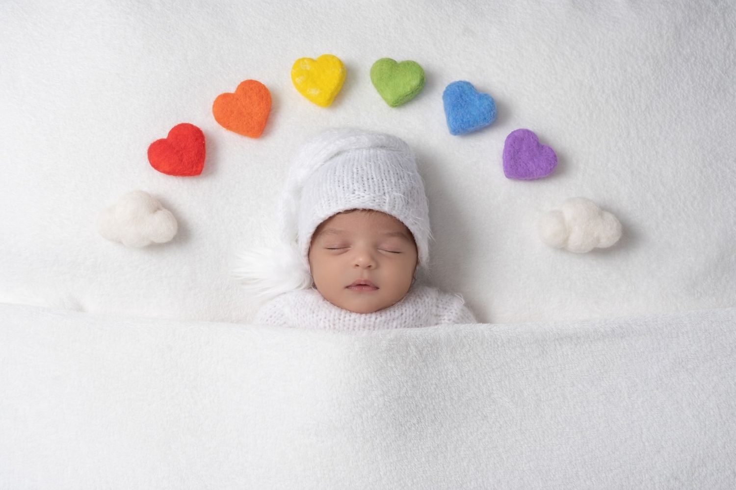 Rainbow colored heart decorations arranged in a row on white background with baby photography setup.