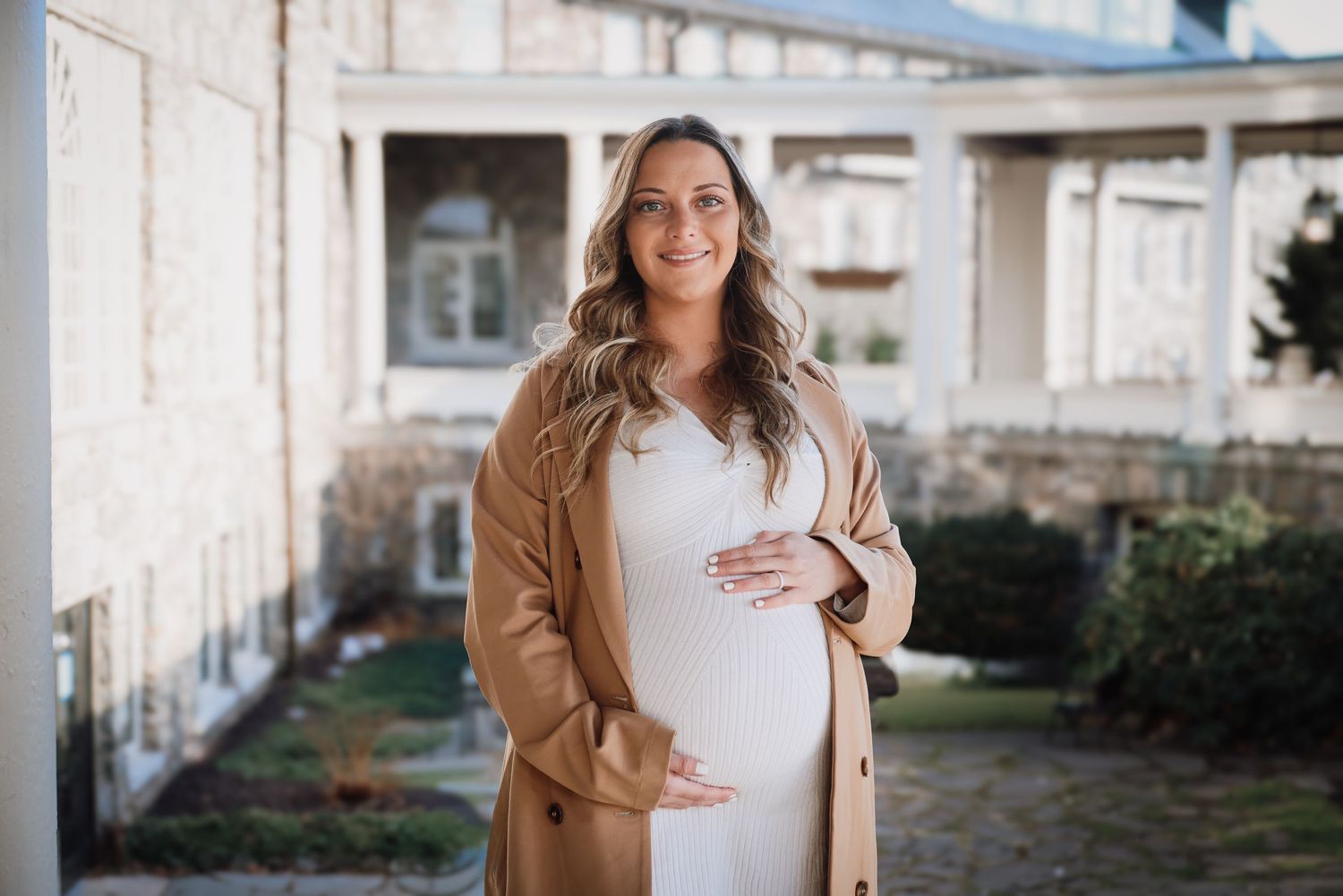 Pregnant woman in white dress and tan cardigan stands outside a building during a maternity photoshoot.
