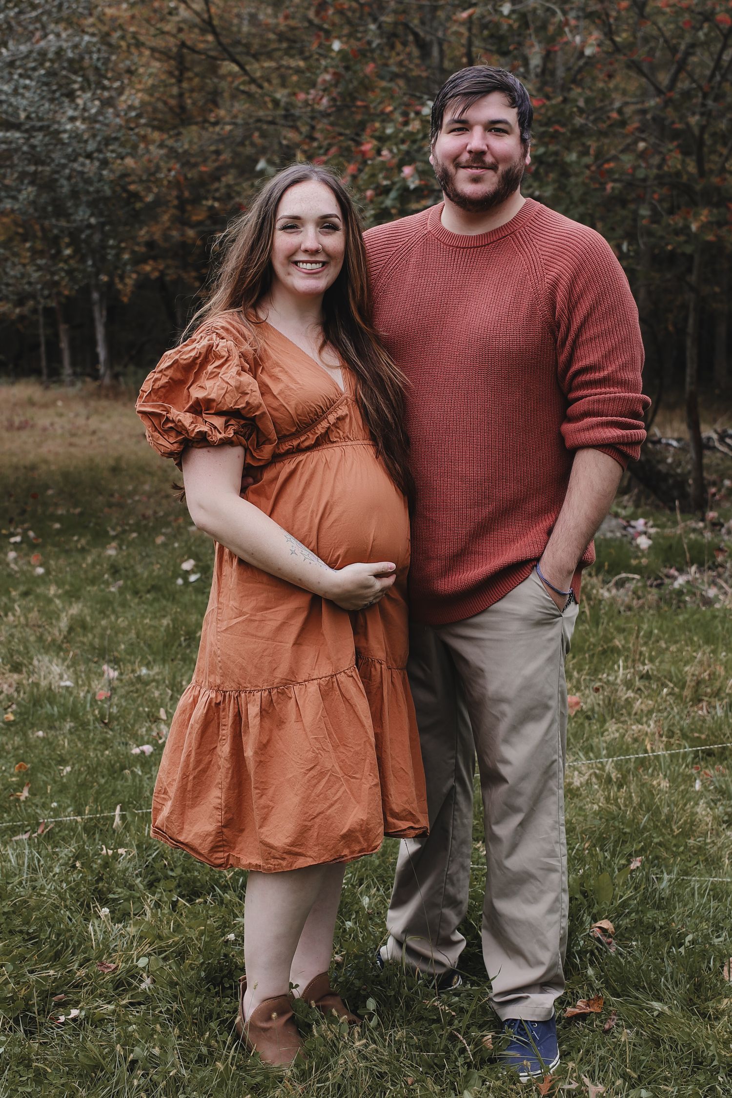 A couple poses outdoors in autumn, wearing rust-colored dress and sweater, with a visible baby bump in a natural setting.