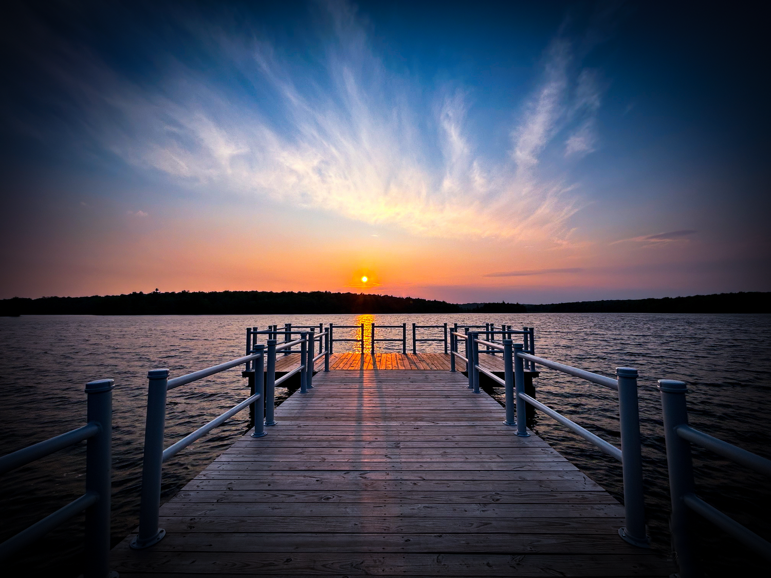 Wooden dock extending over calm lake waters at sunset with dramatic blue sky and radiating clouds.