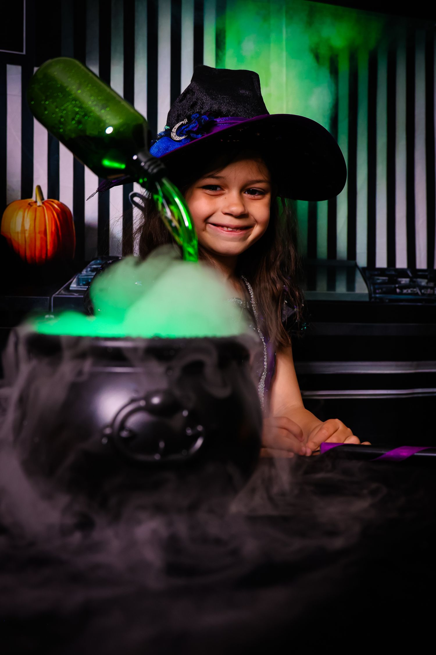 Smiling girl in witch hat poses with smoking Halloween cauldron and green potion bottle in spooky setting.