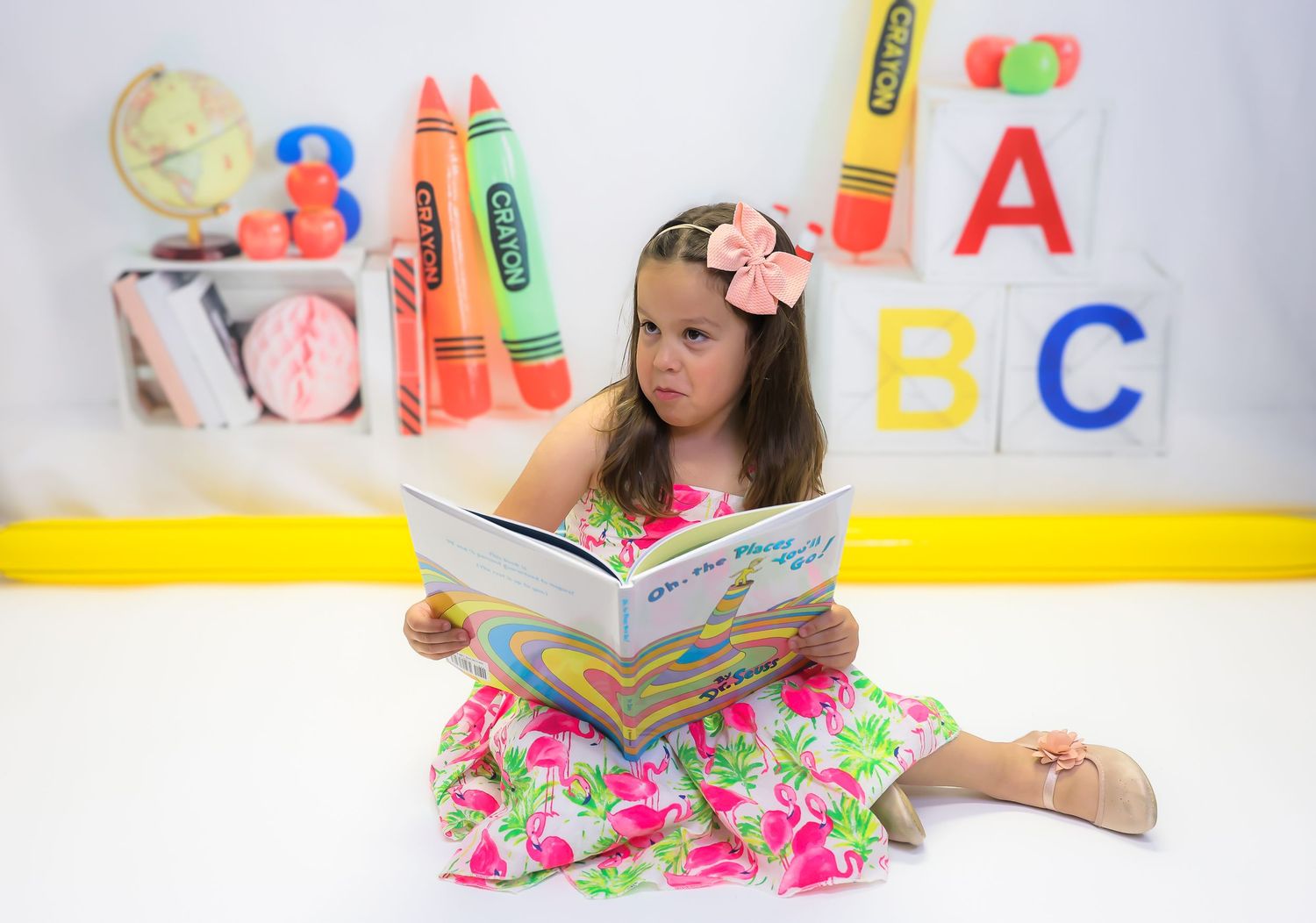 Young girl in floral dress reads a book in a colorful classroom with ABC blocks and crayon decorations.