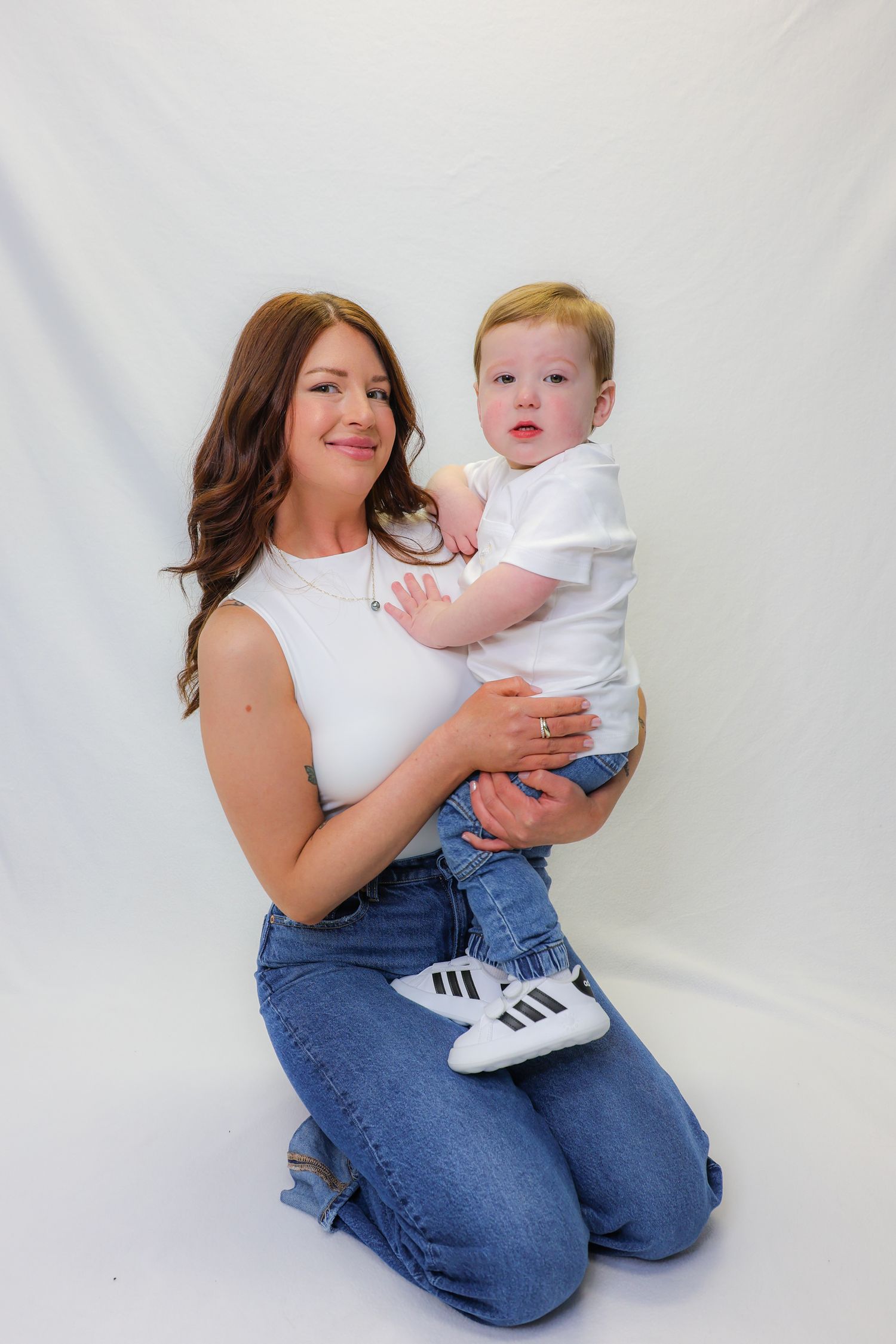 A young woman in white top and jeans kneels holding a baby boy in matching white outfit with Adidas shoes on white backdrop.