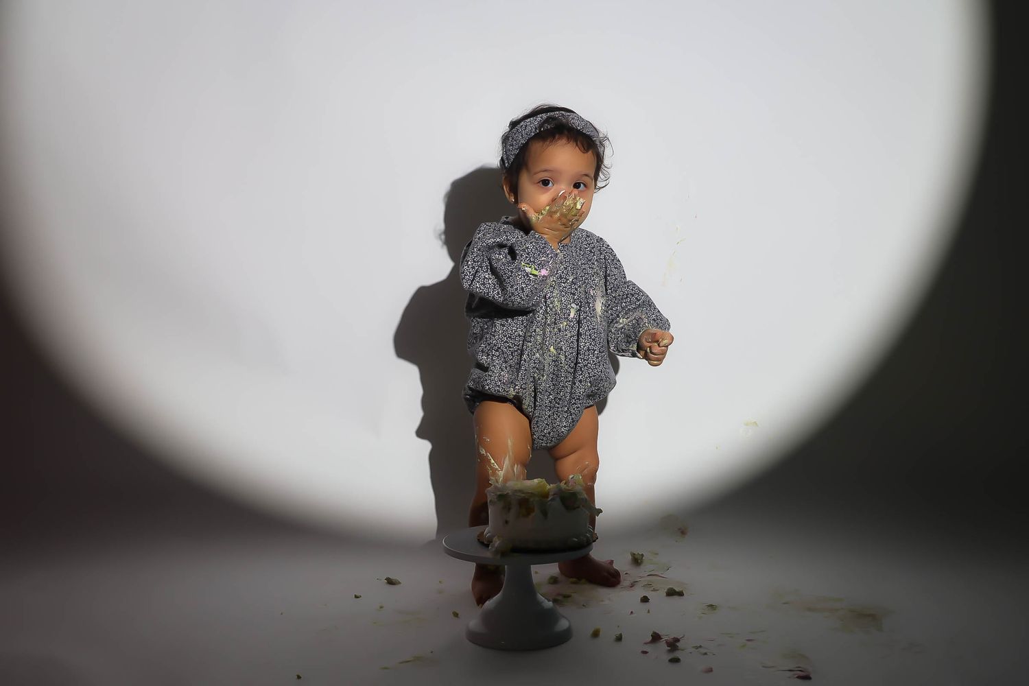 Close-up of a toddler in a gray romper standing over a smashed cake under a circular spotlight.