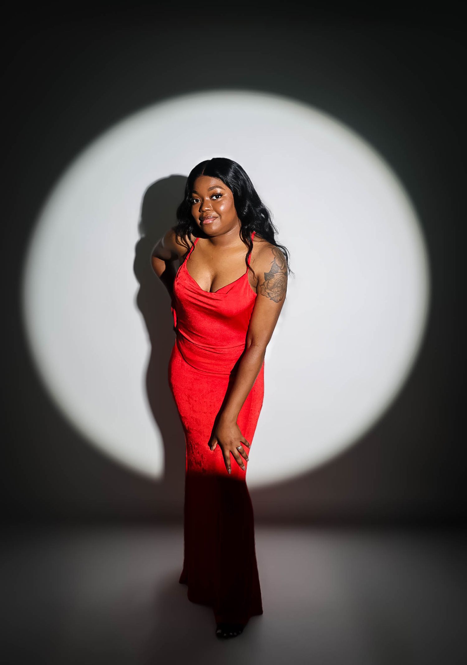 Woman in red gown poses elegantly in spotlight against dark background, showcasing tattoo and long black hair.