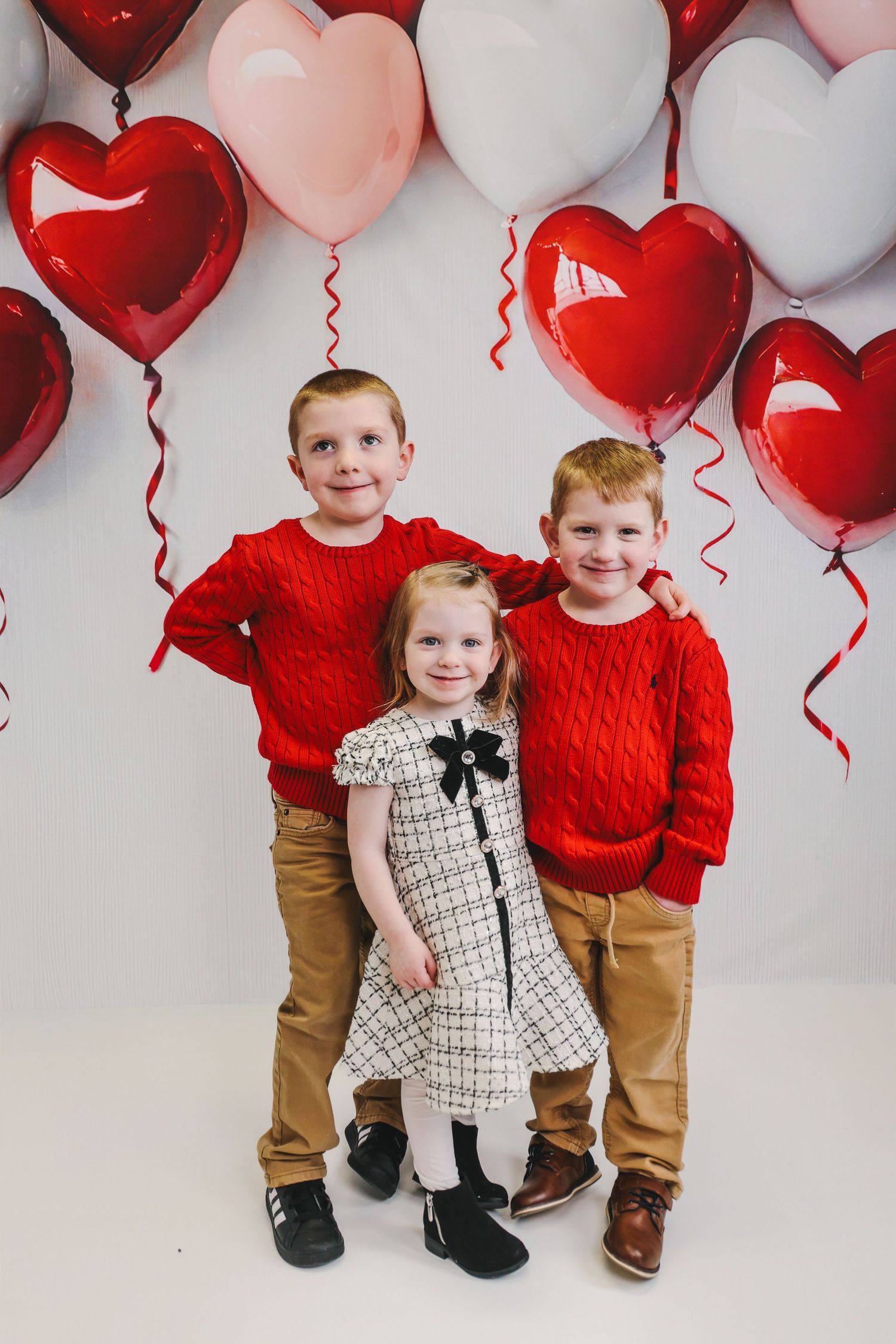 Three children pose with red and pink heart balloons for a Valentine's Day photo, two boys in red sweaters and a girl in a dress.