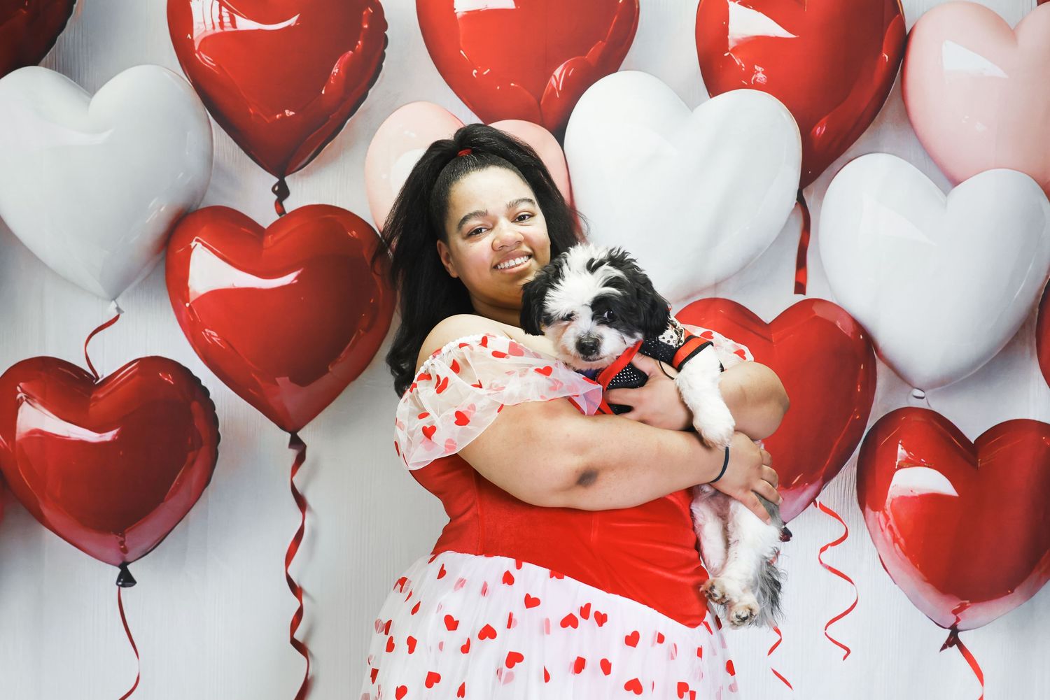 Woman smiling and hugging a small dog surrounded by red and pink heart-shaped balloons on Valentine's Day.