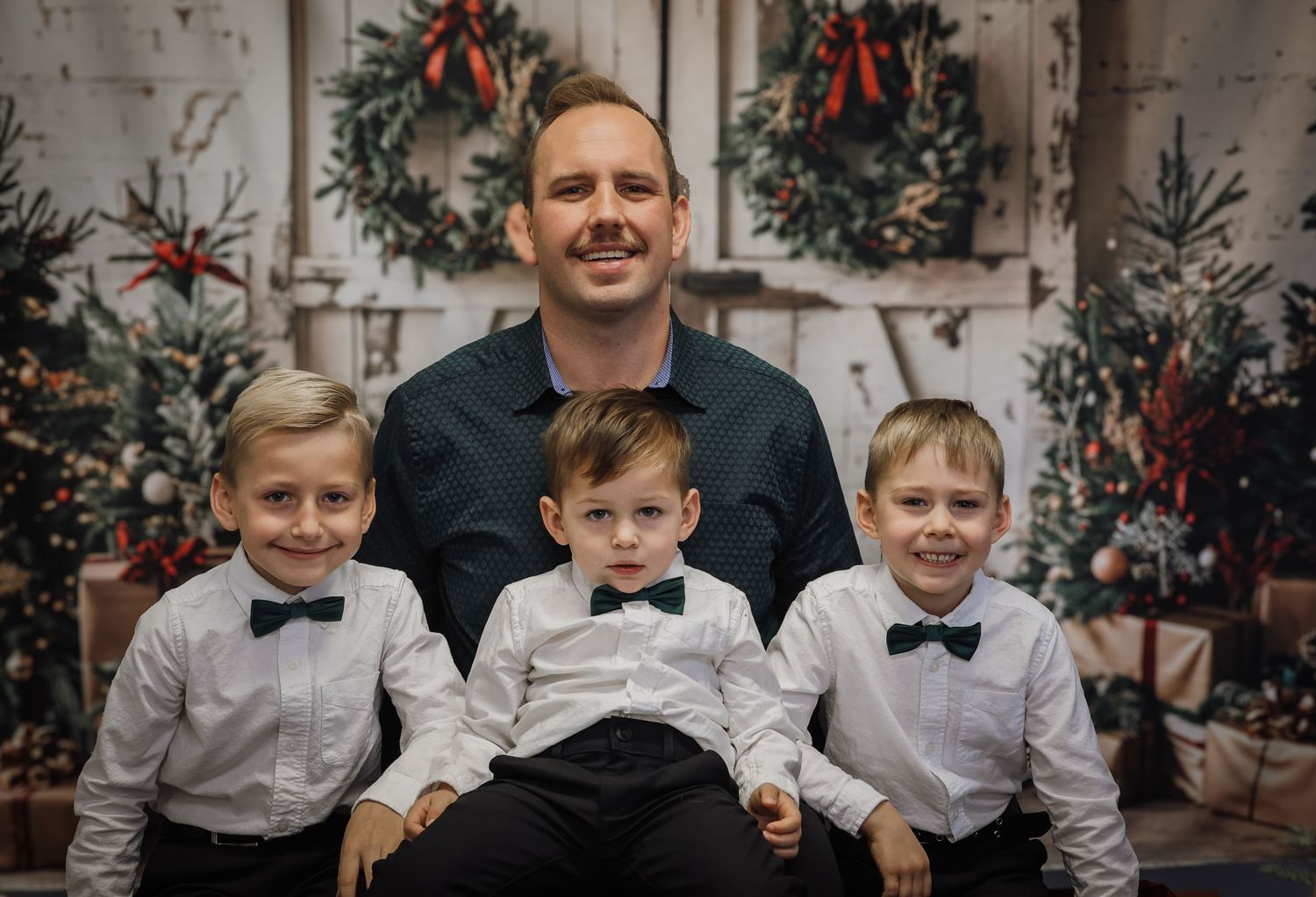 Family Christmas photo with father and three young boys in white shirts and bow ties posed before festive holiday backdrop.