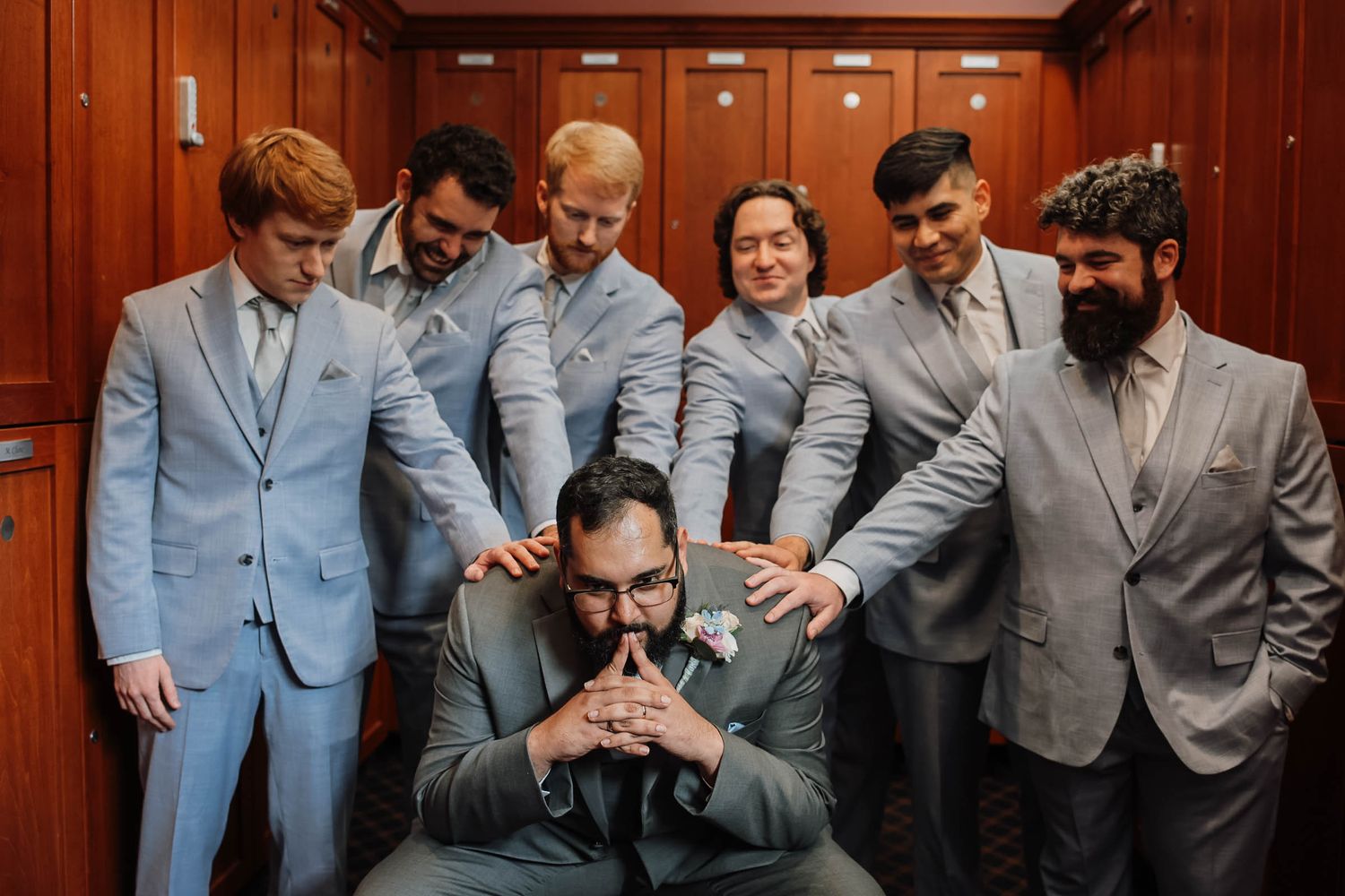Groom seated surrounded by groomsmen in matching light blue suits, sharing a joyful moment in a wood-paneled room.