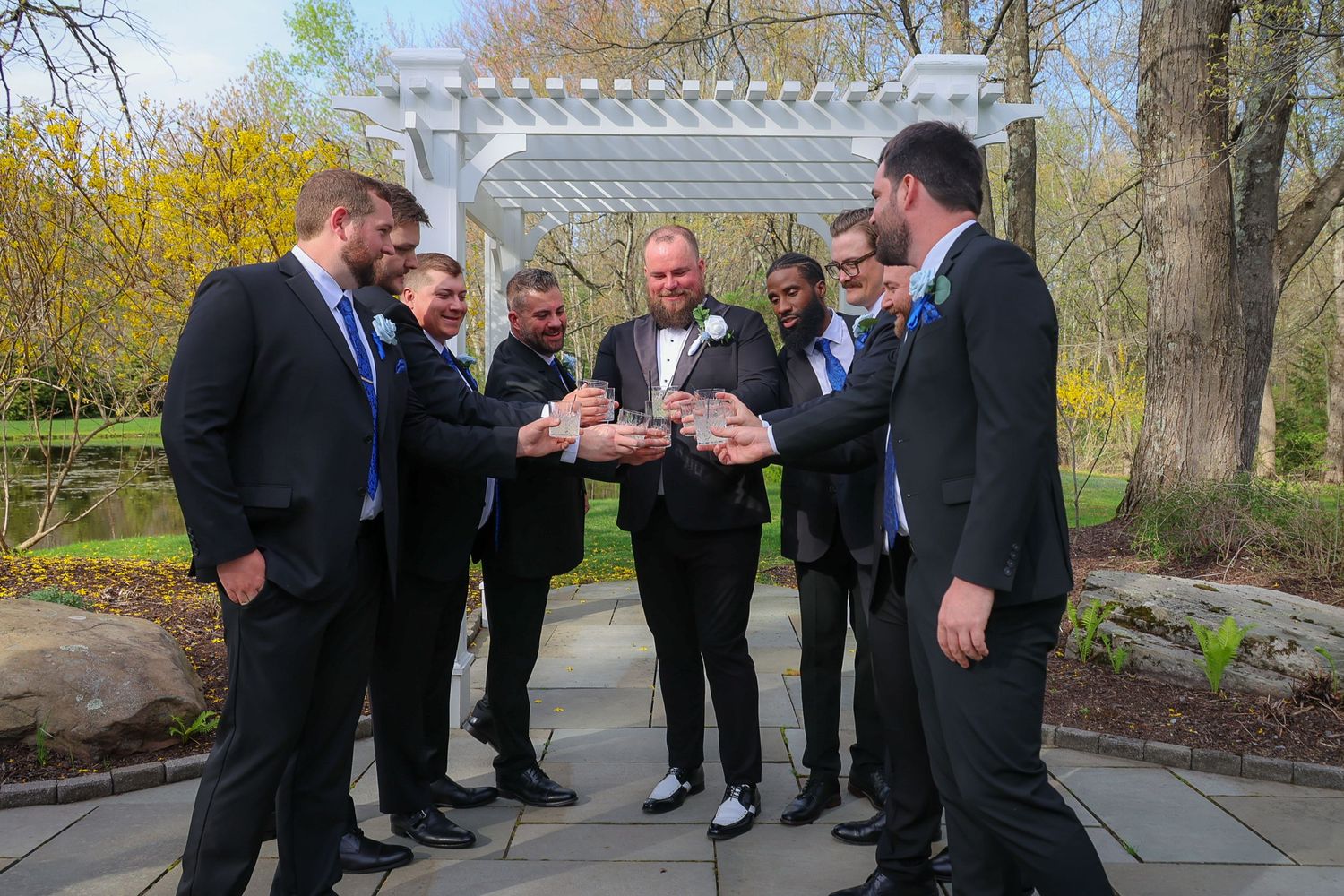 Groom surrounded by groomsmen in black suits examining wedding details in an outdoor garden ceremony venue.