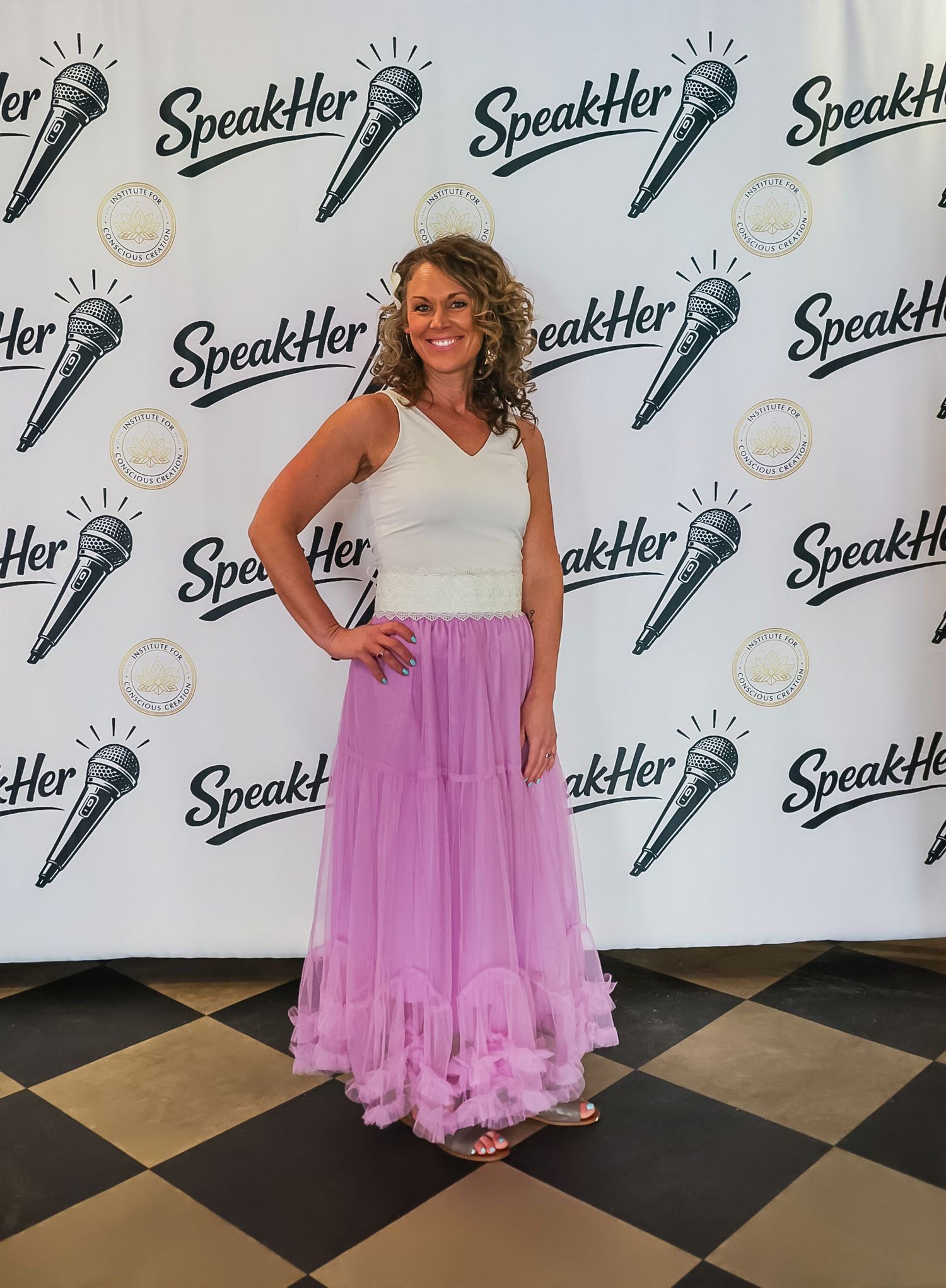 Woman in white top and purple tulle skirt poses at SpeakHer event backdrop.