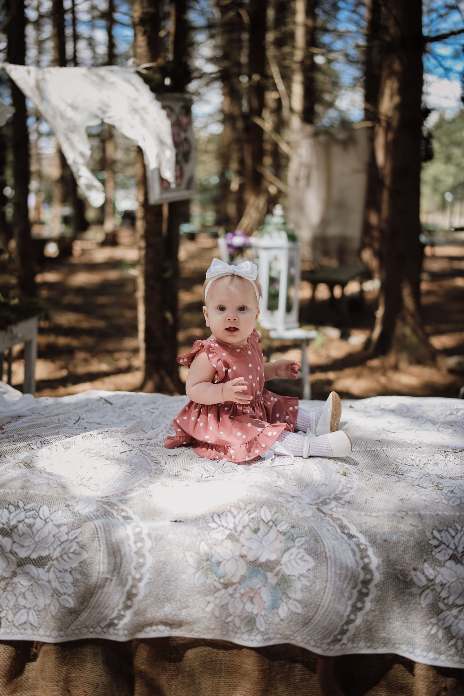 Baby girl in pink polka dot dress sitting on floral blanket outdoors in a sunlit forest setting.