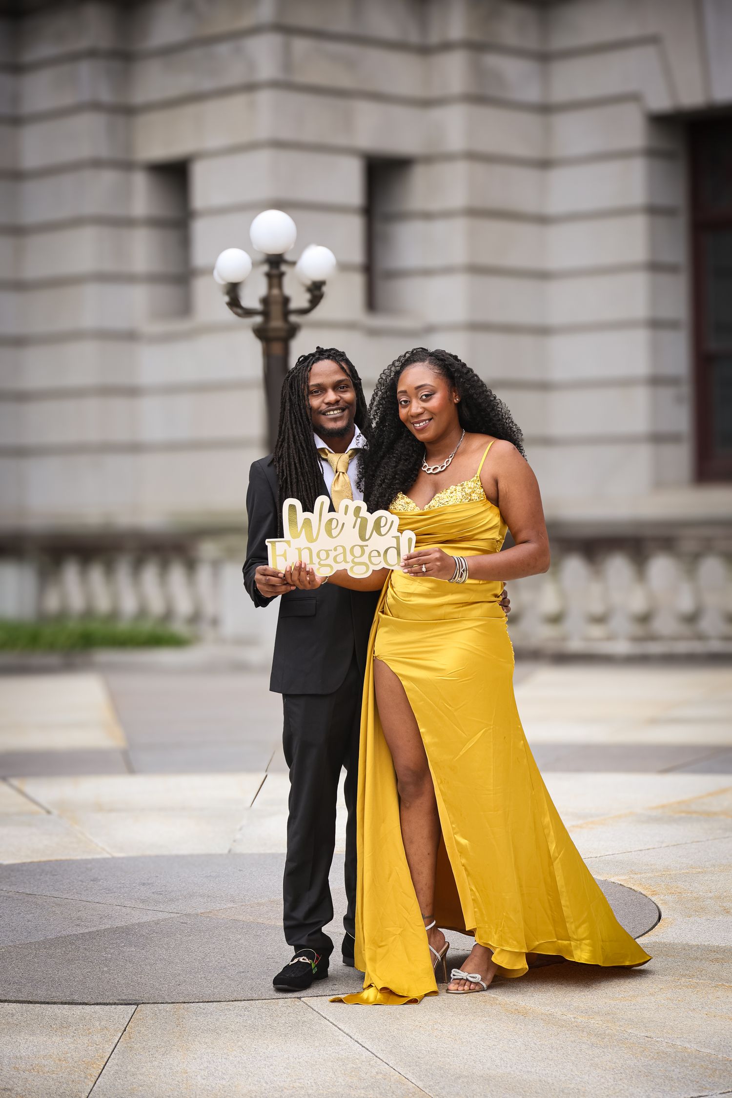 A couple in formal attire poses with a decorative 'Happy Birthday' sign outside a grand stone building.