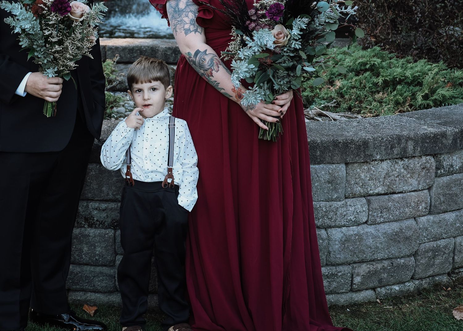 A young child in vintage-style clothing with suspenders stands beside a burgundy dress against a stone wall in a garden setting.
