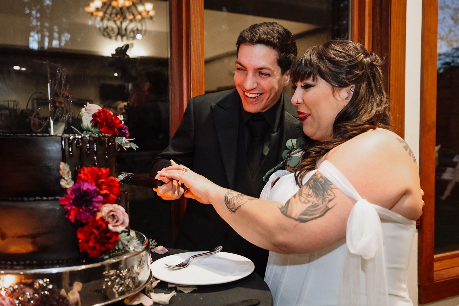 A couple shares a joyful moment while cutting their wedding cake together at an indoor reception.