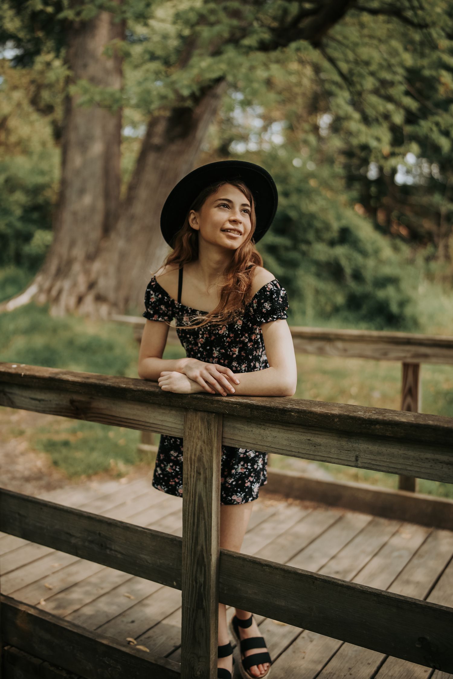 A person in a floral dress and hat leans on a wooden railing in a park setting during golden hour.