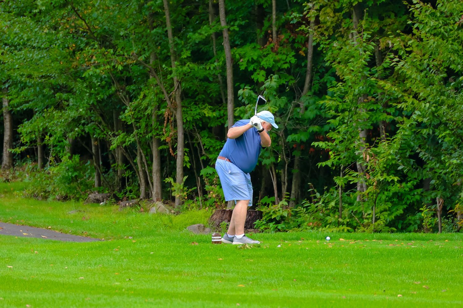A golfer takes a swing on a lush green golf course surrounded by trees on a sunny day.