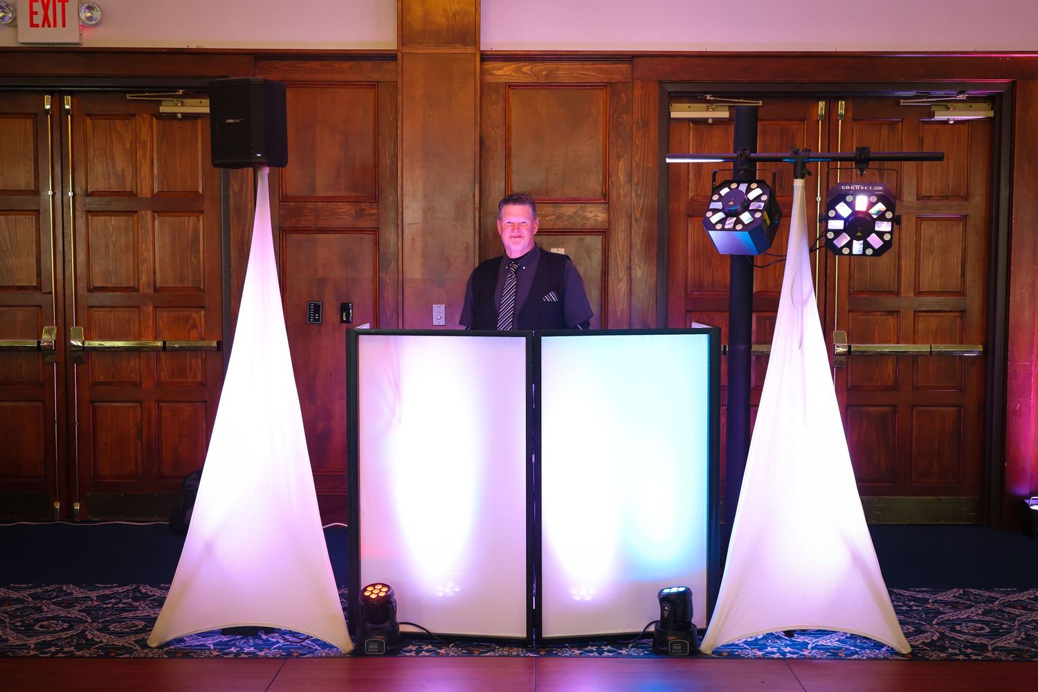 A DJ booth setup with illuminated white panels and decorative triangular light towers at a formal event venue.