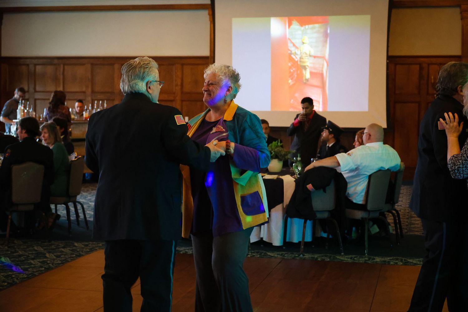 Two people dance together at an indoor event venue with tables and guests visible in the background.