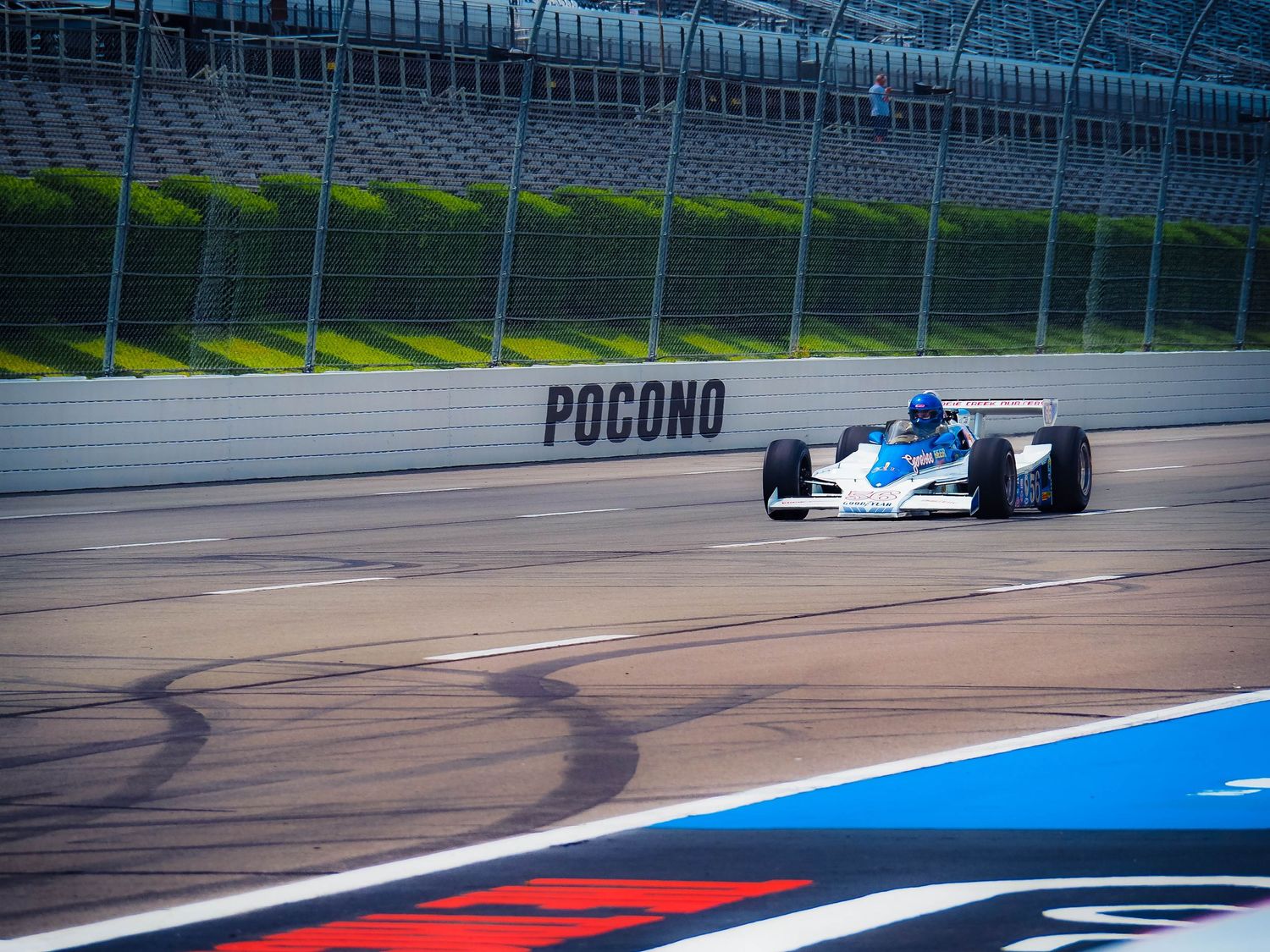 Race car speeds around turn at Pocono Raceway leaving tire marks on blue and red painted track surface.
