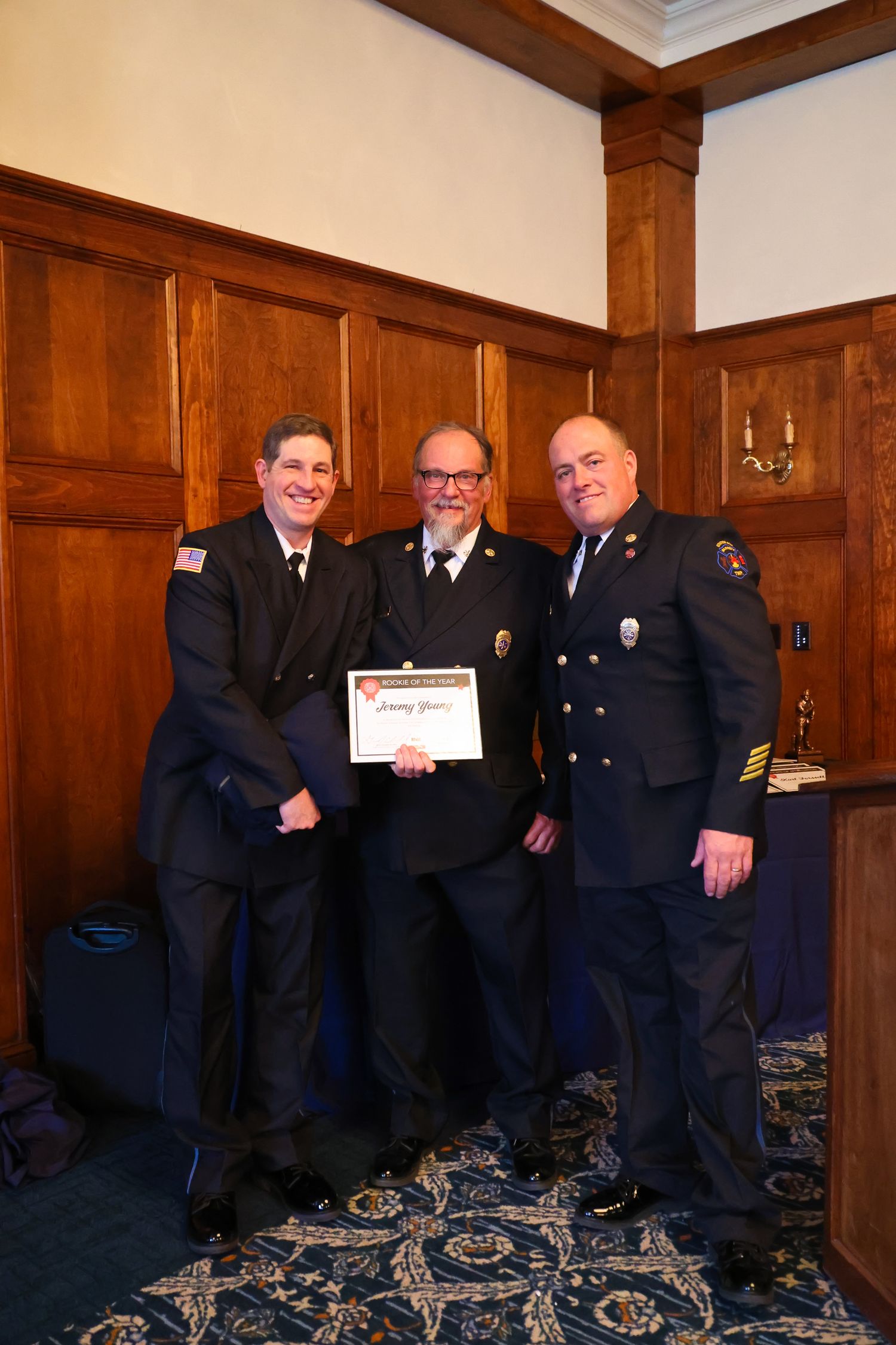 A group of uniformed fire officials stand together in a wood-paneled room with ornate blue carpeting.