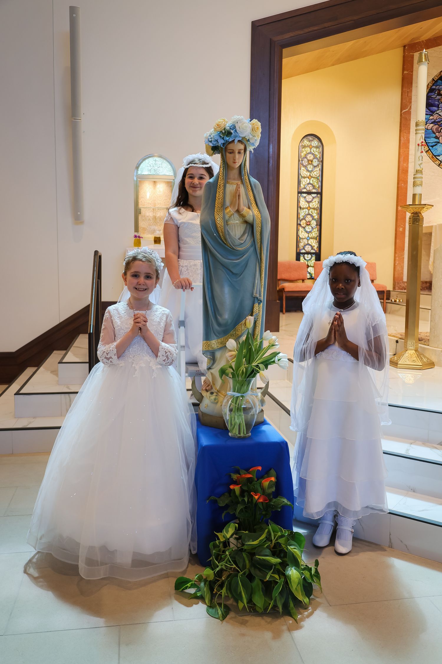 Children in white First Communion dresses kneel in prayer at a church altar beside a religious statue.