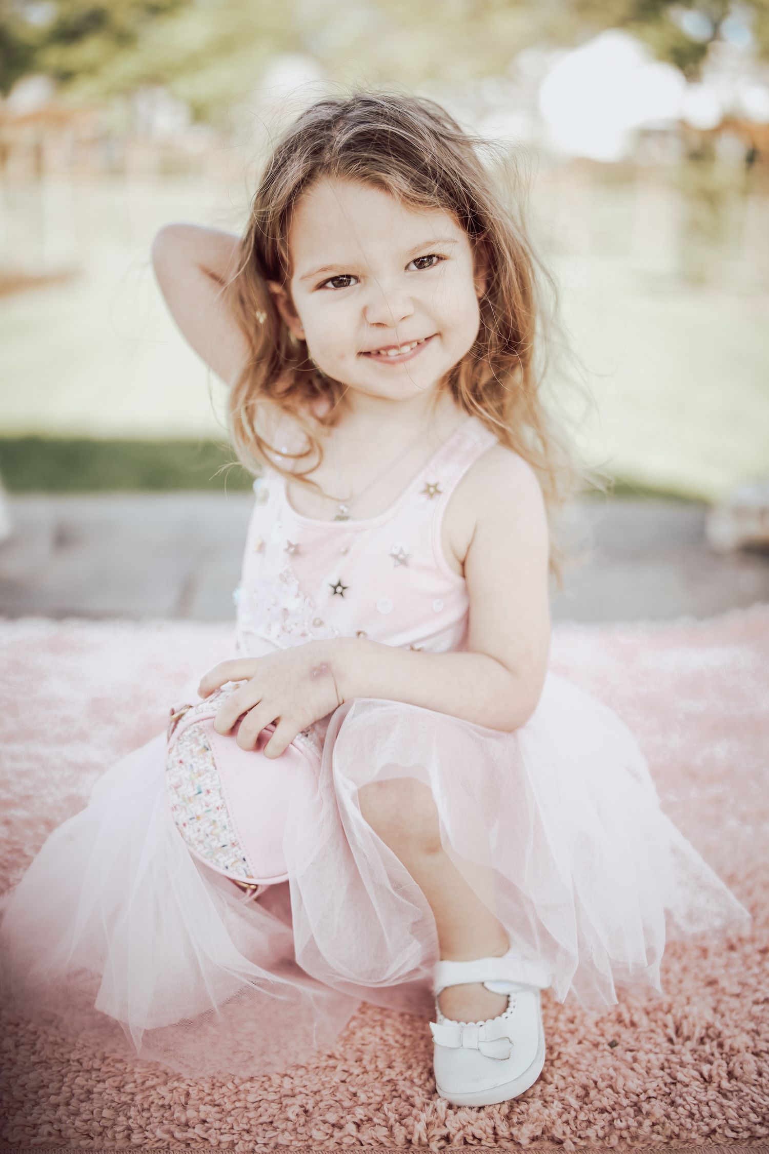 A young child in a pink tulle dress and white shoes poses outdoors in soft lighting.