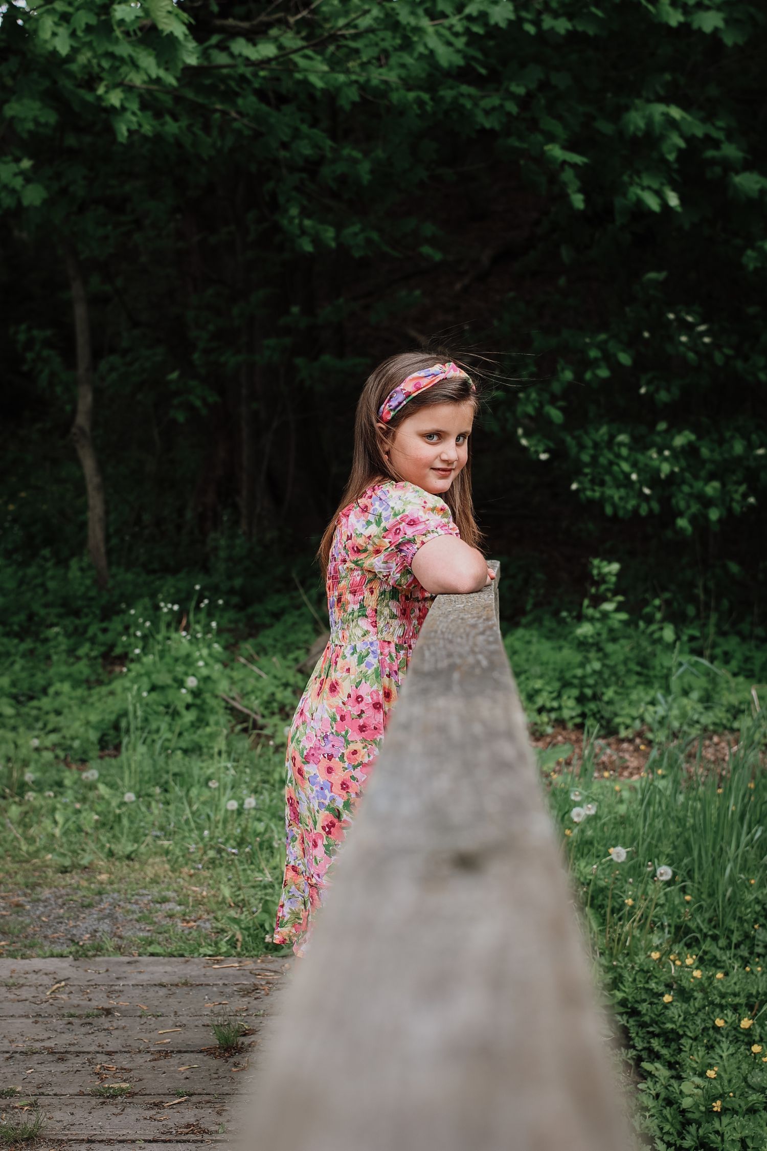 A person in a floral dress poses playfully in a wooded area with lush greenery and wildflowers.