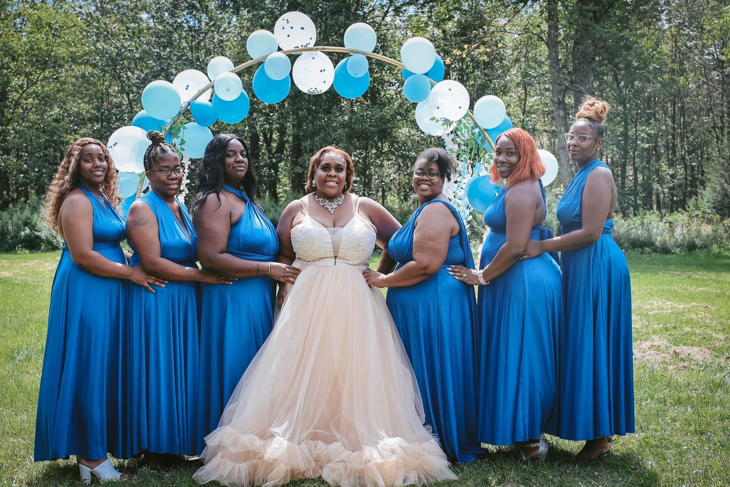 A wedding party poses under a blue and white balloon arch outdoors, with bridesmaids in bright blue dresses and a bride in blush.