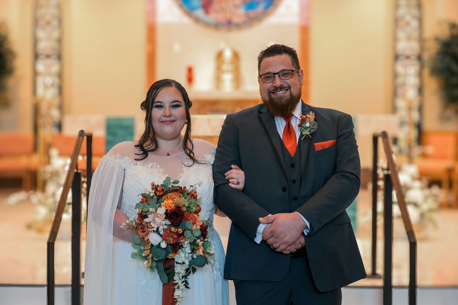 A couple stands at the altar during their wedding ceremony, with orange and white floral bouquet and decorative details.