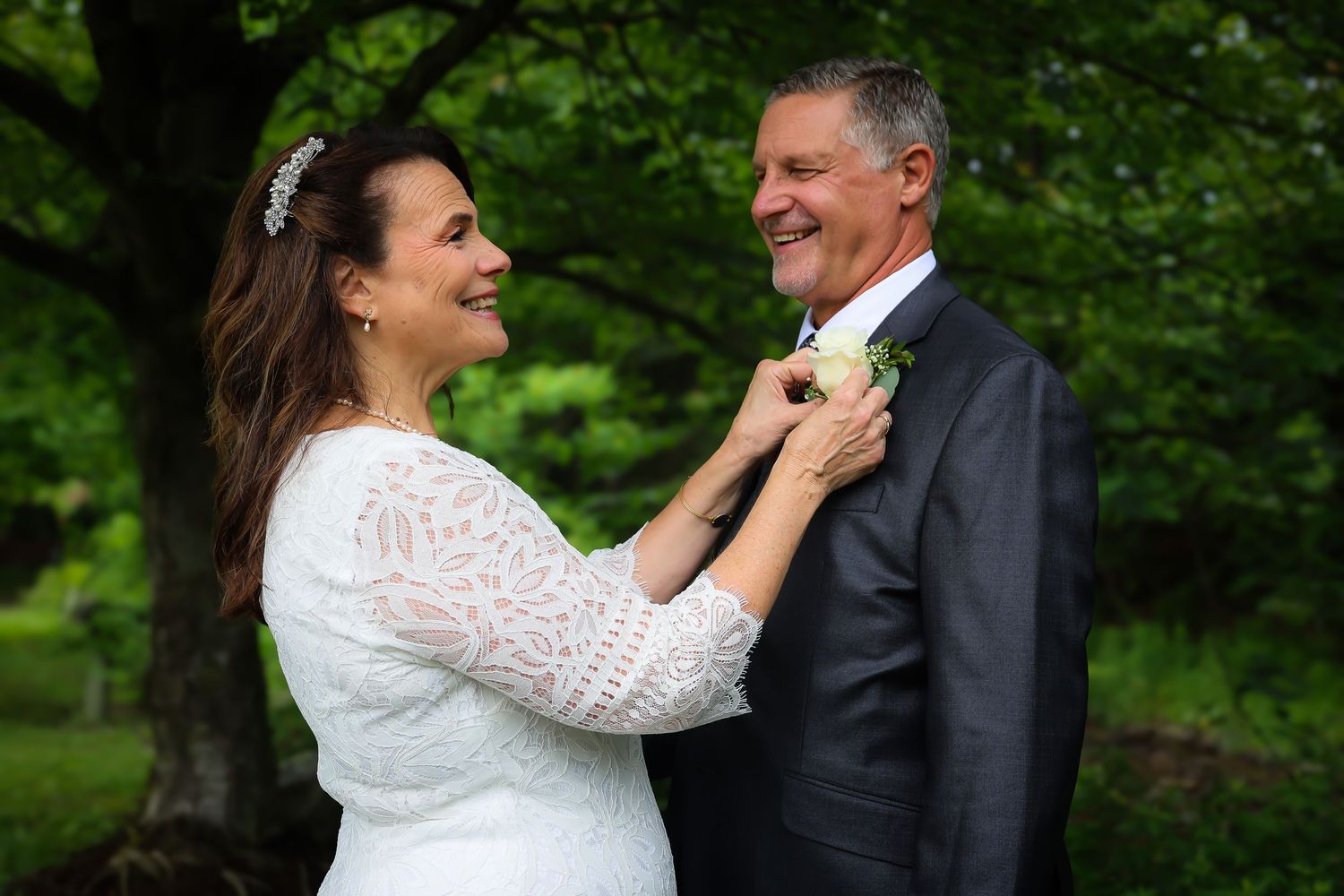 A couple shares a joyful moment outdoors as the bride adjusts the groom's boutonniere against a lush green background.