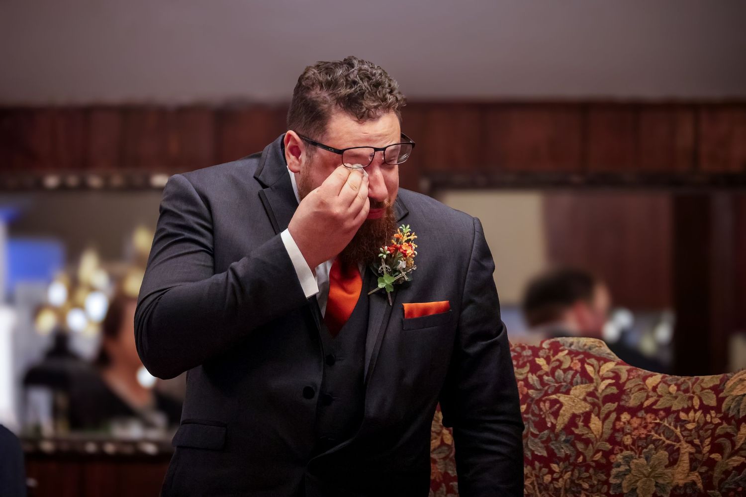 An emotional groom in a black suit with orange boutonniere wipes his eyes during a wedding ceremony.