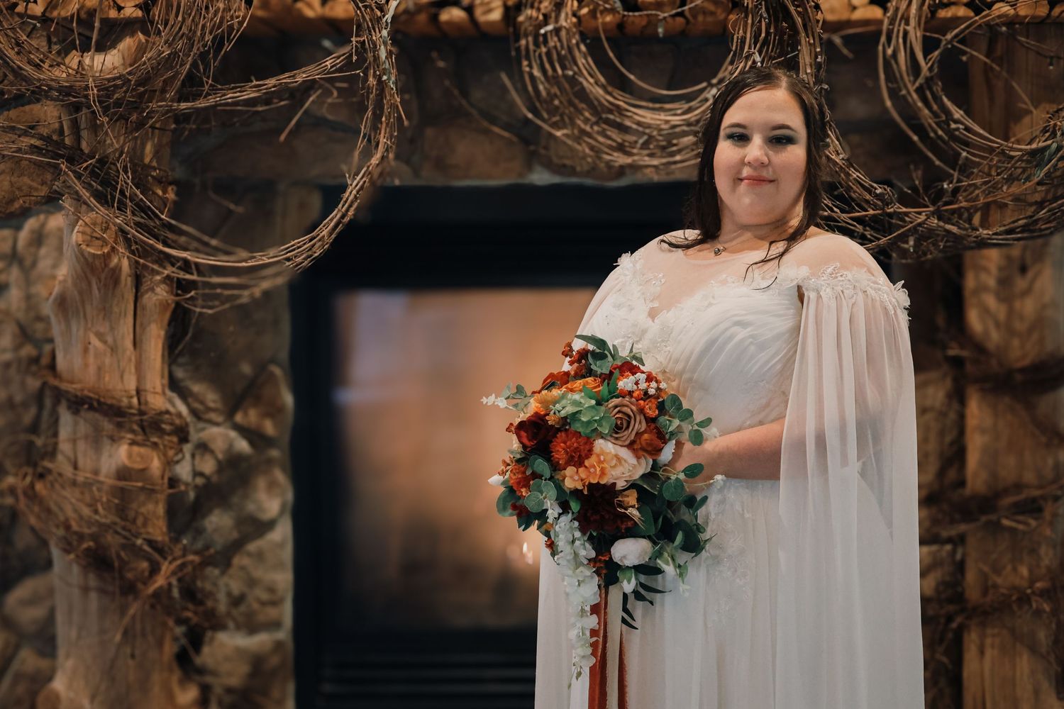 A bride in a flowing white dress holds a vibrant red and white bouquet in a rustic outdoor setting.